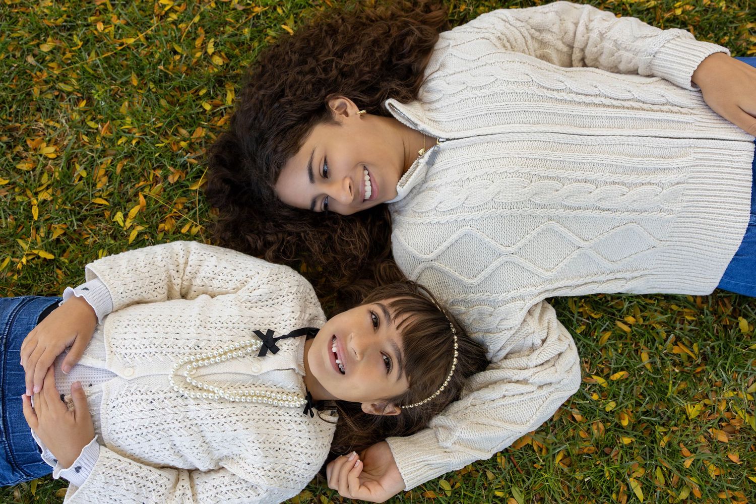 Two girls in cream sweaters lying on green grass covered with autumn leaves, smiling and enjoying outdoor time together.