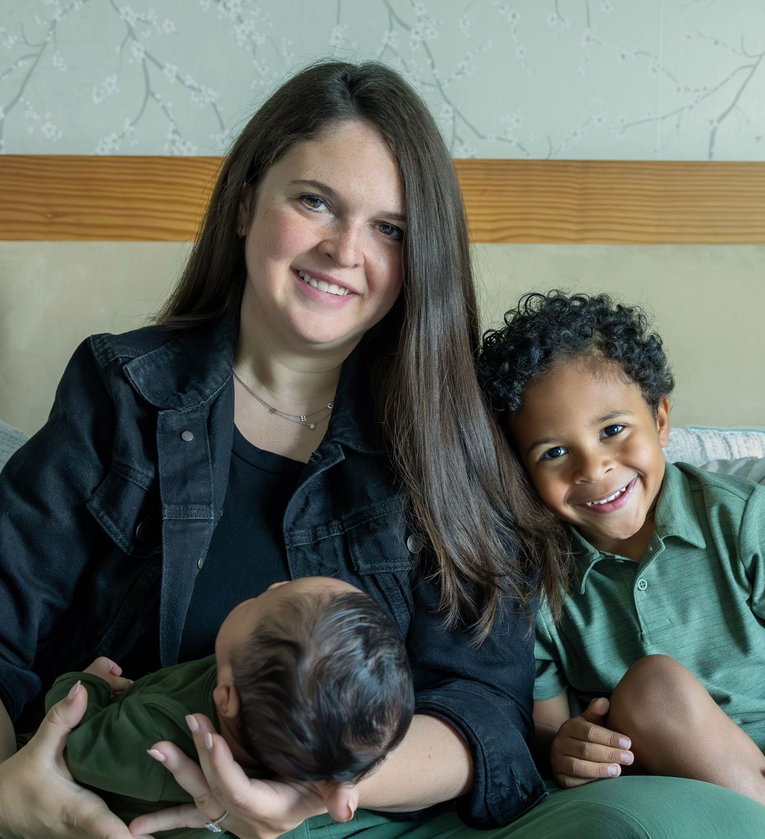 A smiling woman holds a newborn baby while sitting with a young curly-haired boy in a green shirt on a bed.