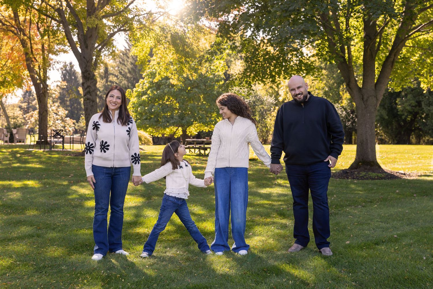 Family of four holding hands and walking in a sunny park with green trees and grass in autumn.