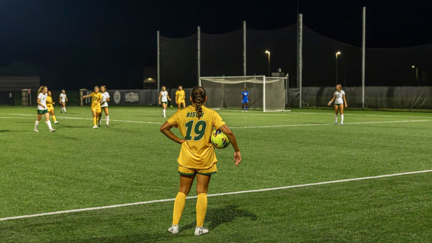 Soccer players in yellow uniforms compete on an illuminated field during a nighttime match.
