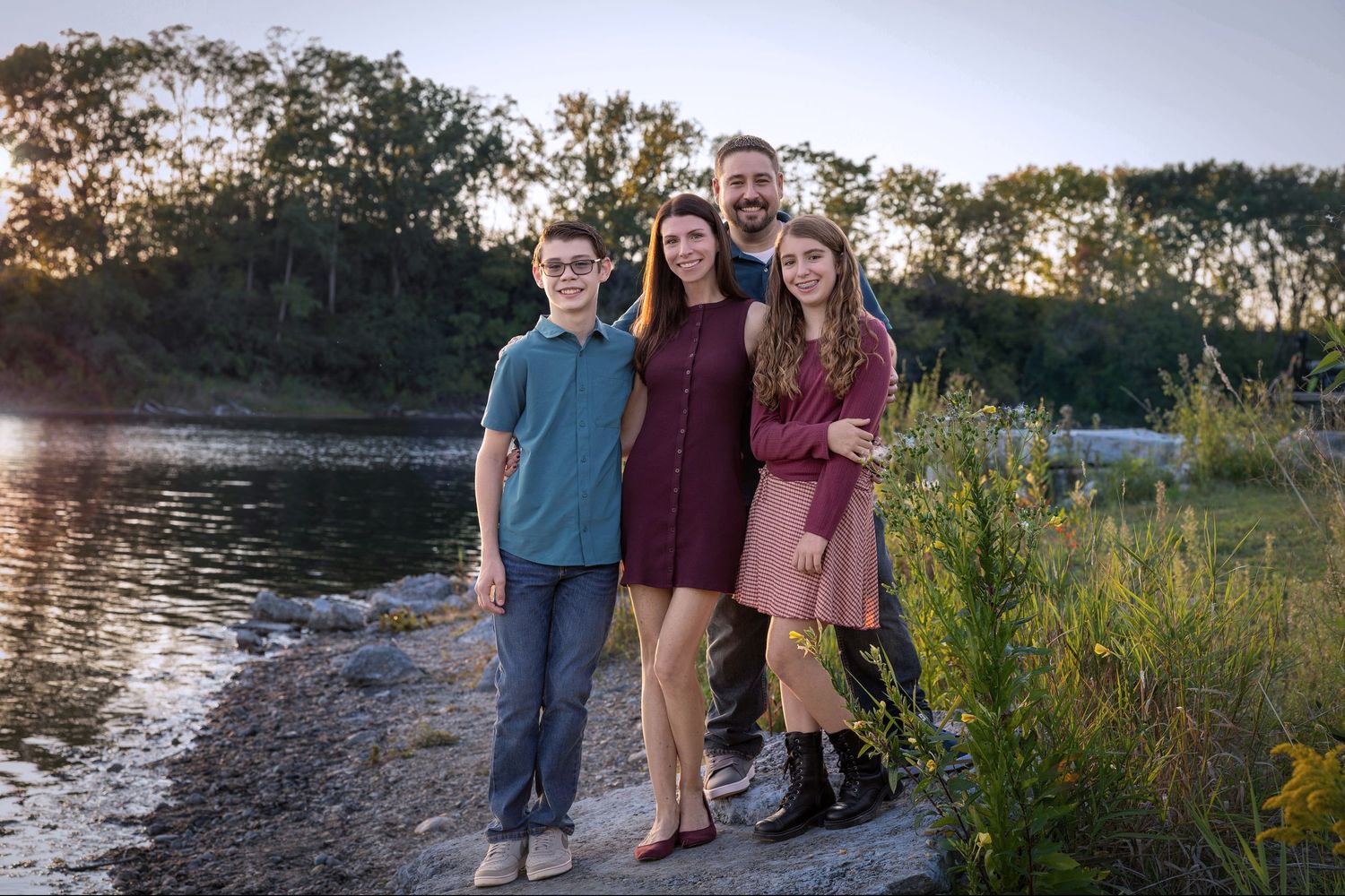 A group stands together on a rocky shoreline at sunset with golden light filtering through trees in the background.