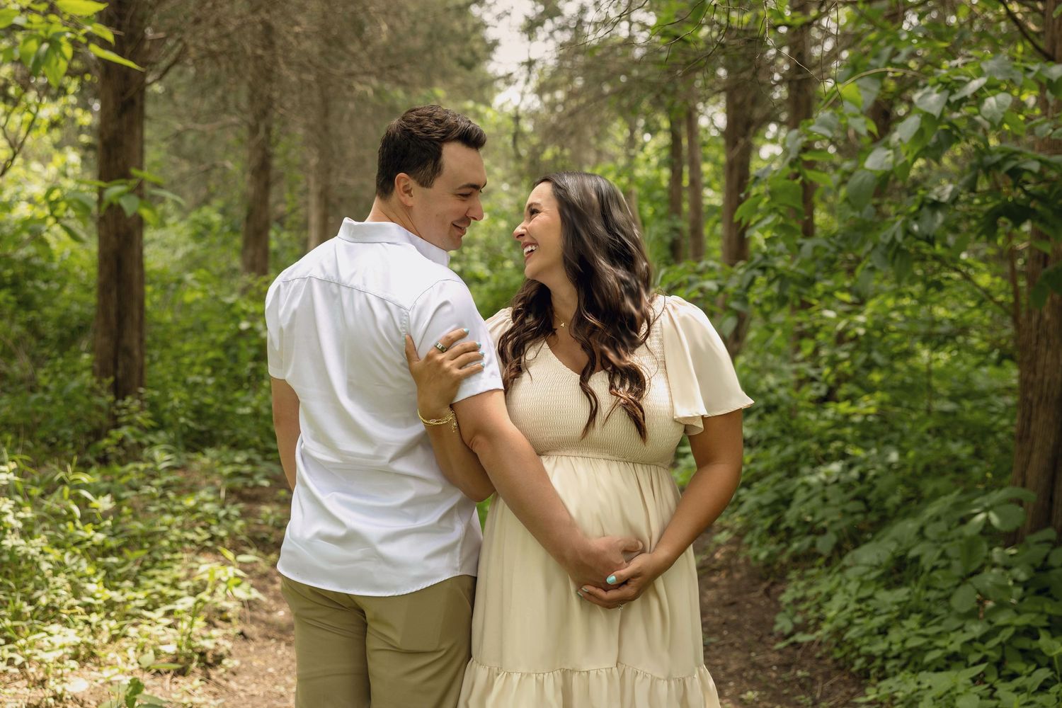 Couple embracing on a forest path, woman in cream maternity dress, man in white shirt, surrounded by lush green trees.
