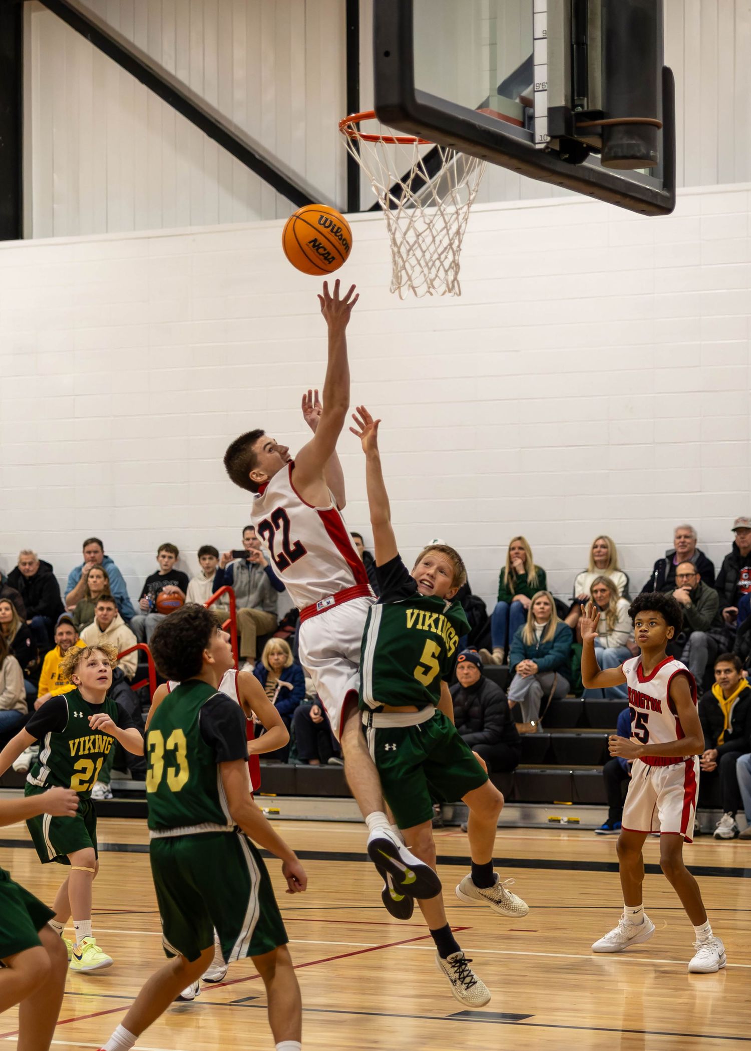 High school basketball player in white uniform attempts a layup over a Vikings defender #5 during an indoor game.