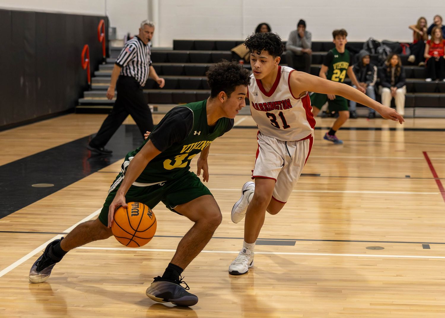 Two high school basketball players compete on indoor court, green uniform player dribbling past red uniform defender.