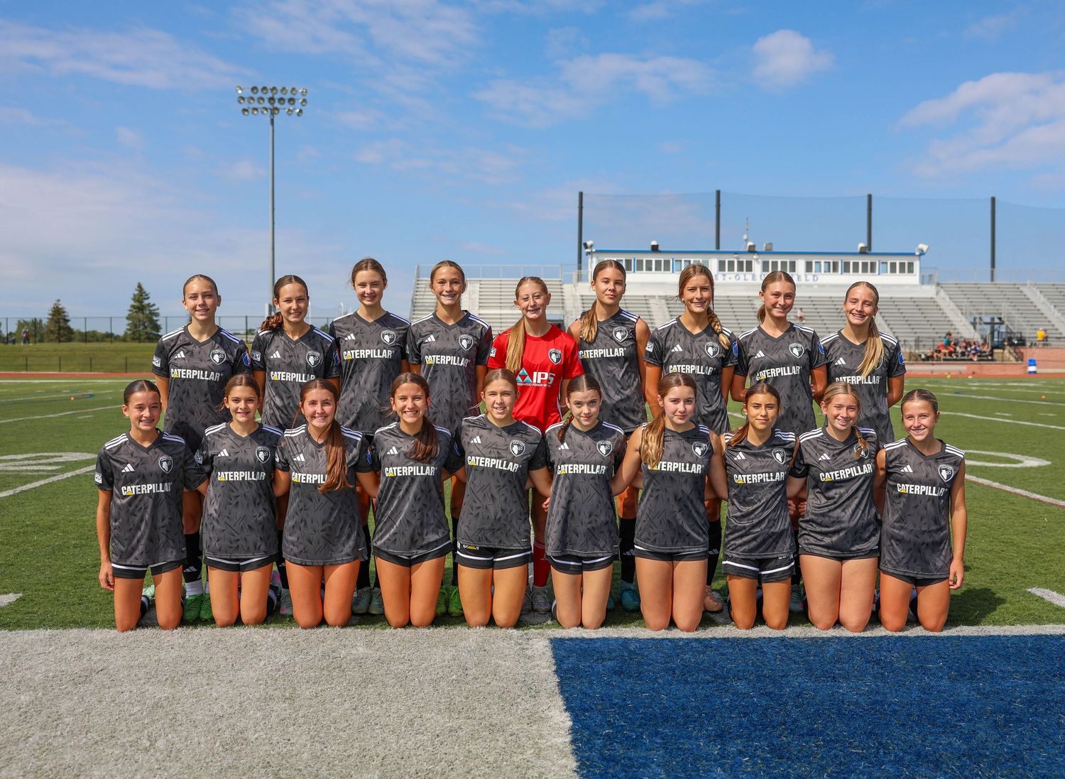 A group of soccer players in matching gray jerseys stand together on a field against a blue sky backdrop.