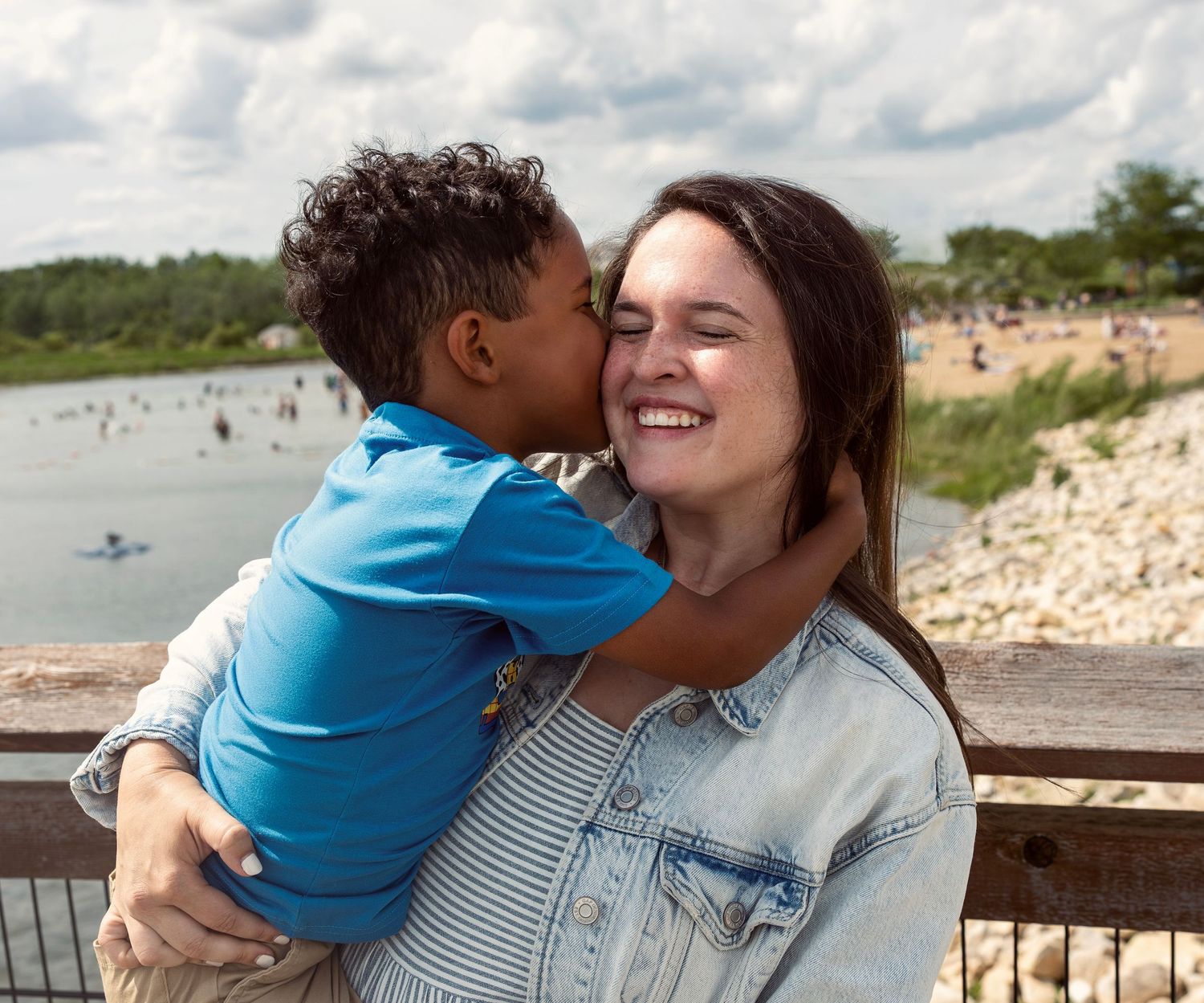A young boy kisses his smiling mother on the cheek outdoors near a lake on a cloudy day.