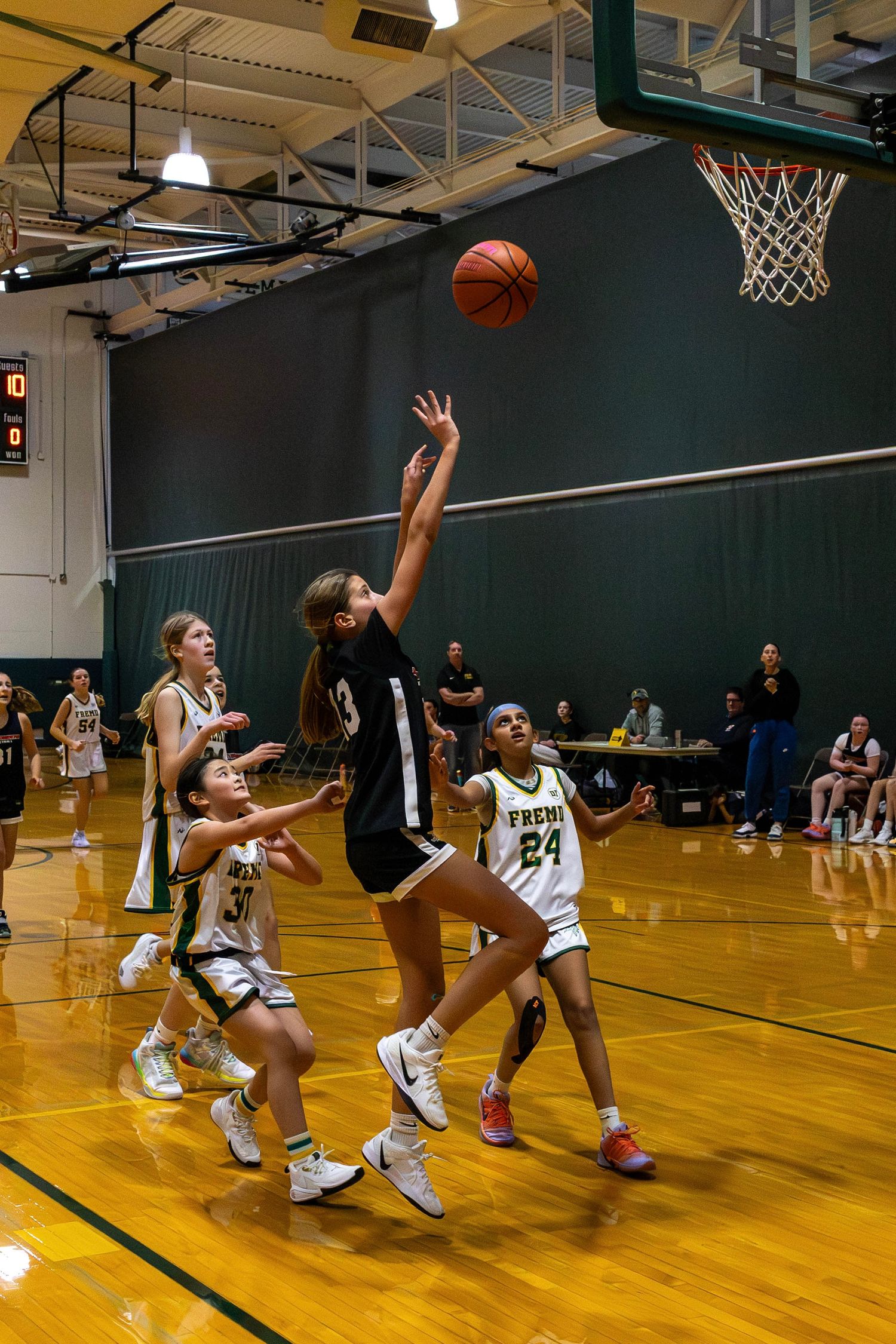 Girls basketball player in black uniform leaps for a layup during an indoor game against green-uniformed opponents.
