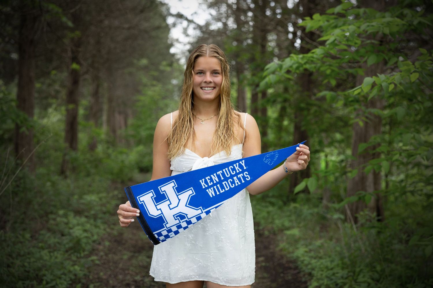Person in white dress holding a blue University of Kentucky spirit banner in a forest setting.