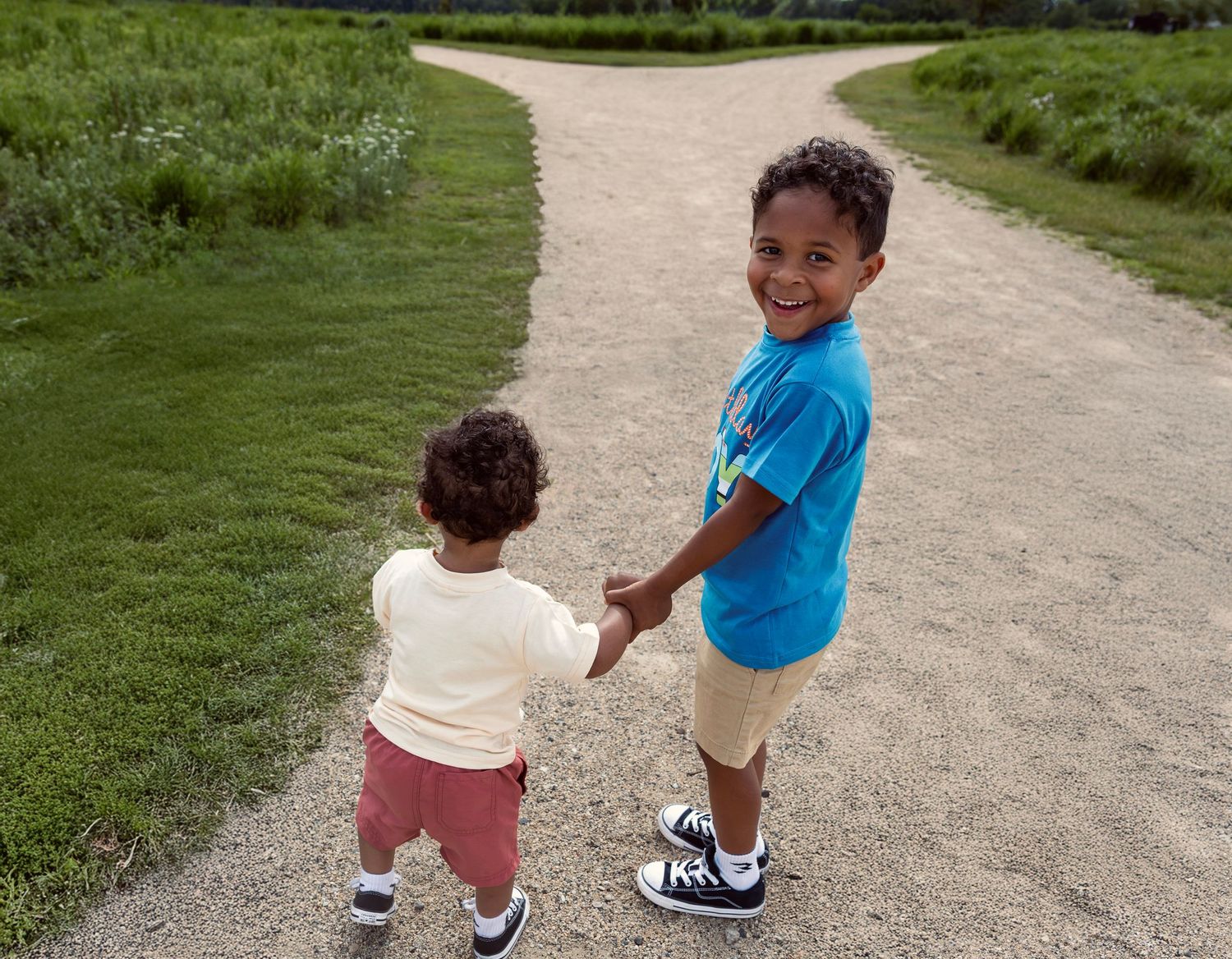 Two young boys holding hands on a gravel path, smiling outdoors surrounded by green grass.