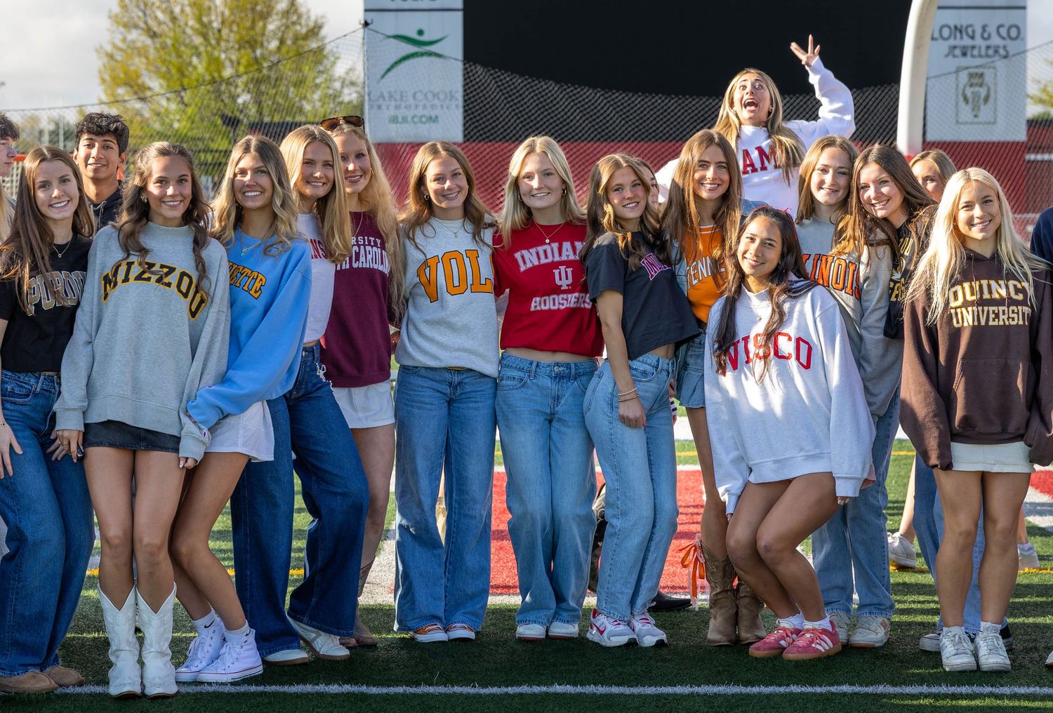 Group of teenage girls posing together outdoors wearing college sweatshirts on a sports field.