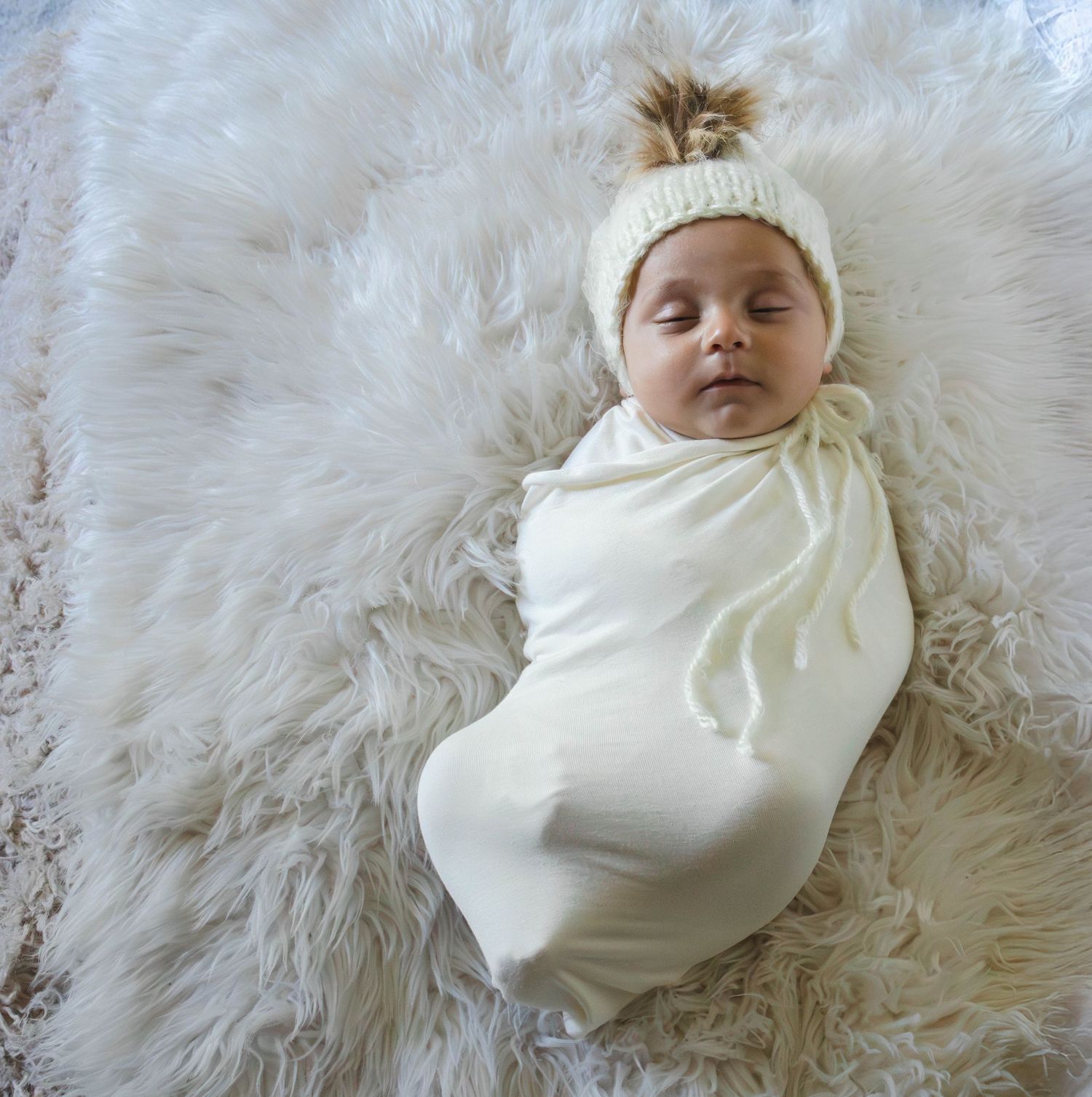 Newborn baby swaddled in cream wrap wearing knit pom-pom hat, lying on fluffy white fur rug.