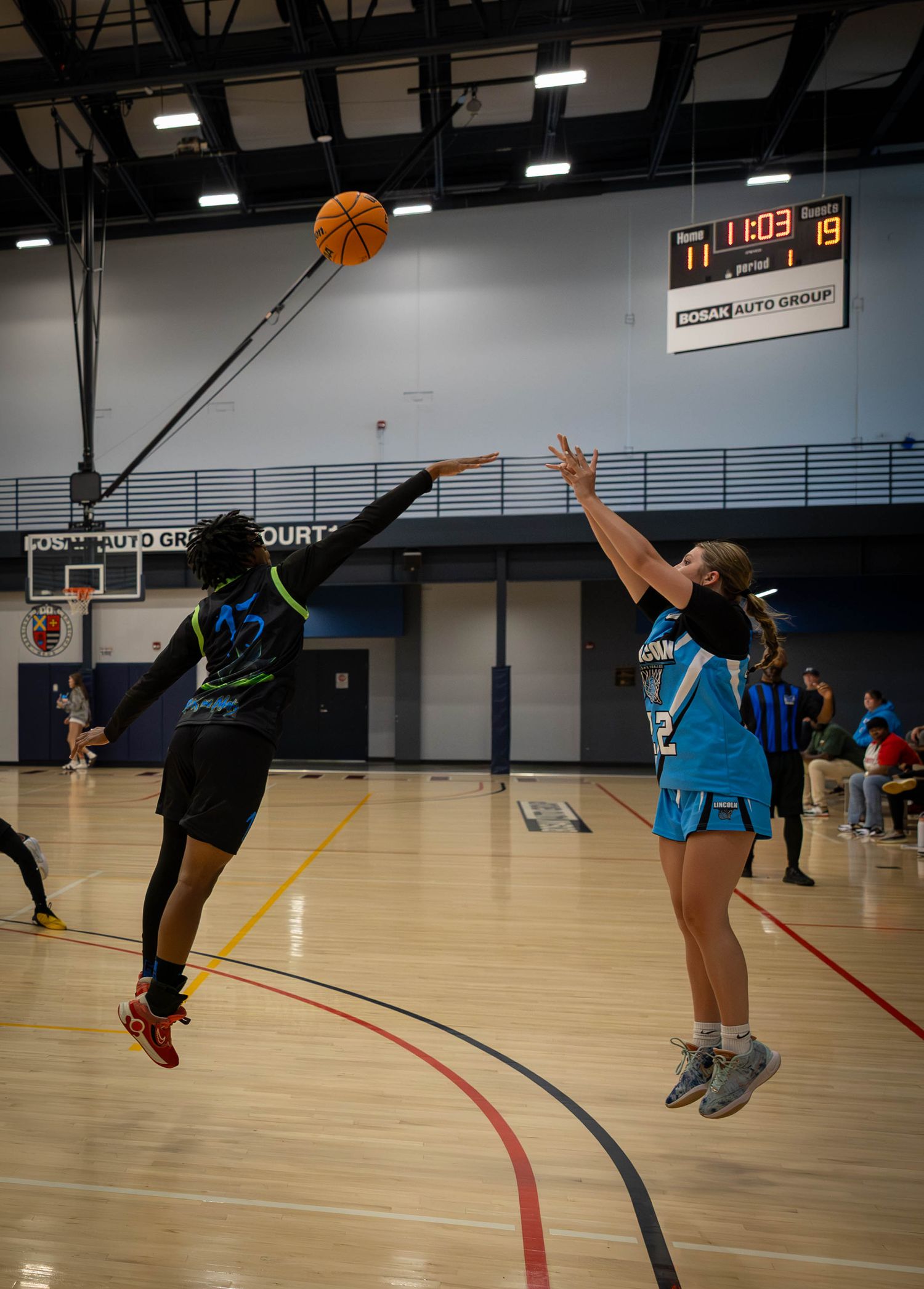 Two basketball players jump for the ball during an indoor game in a gymnasium with a scoreboard visible.