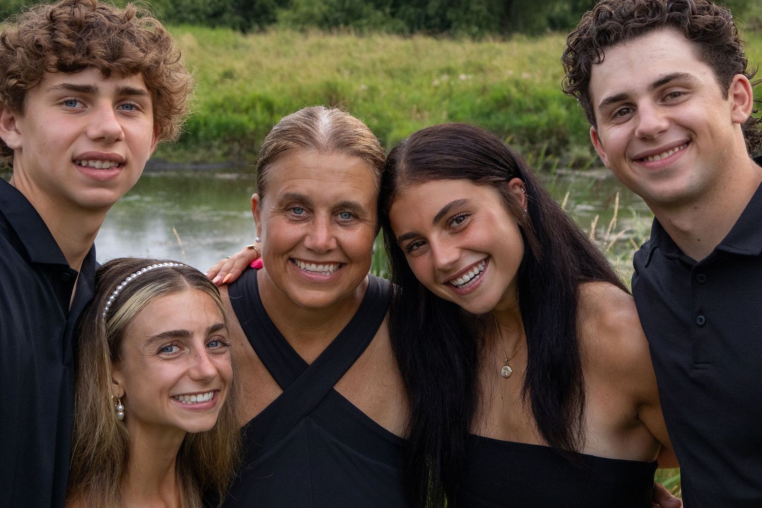 Five smiling young adults in black outfits pose together outdoors near a pond surrounded by lush green trees.