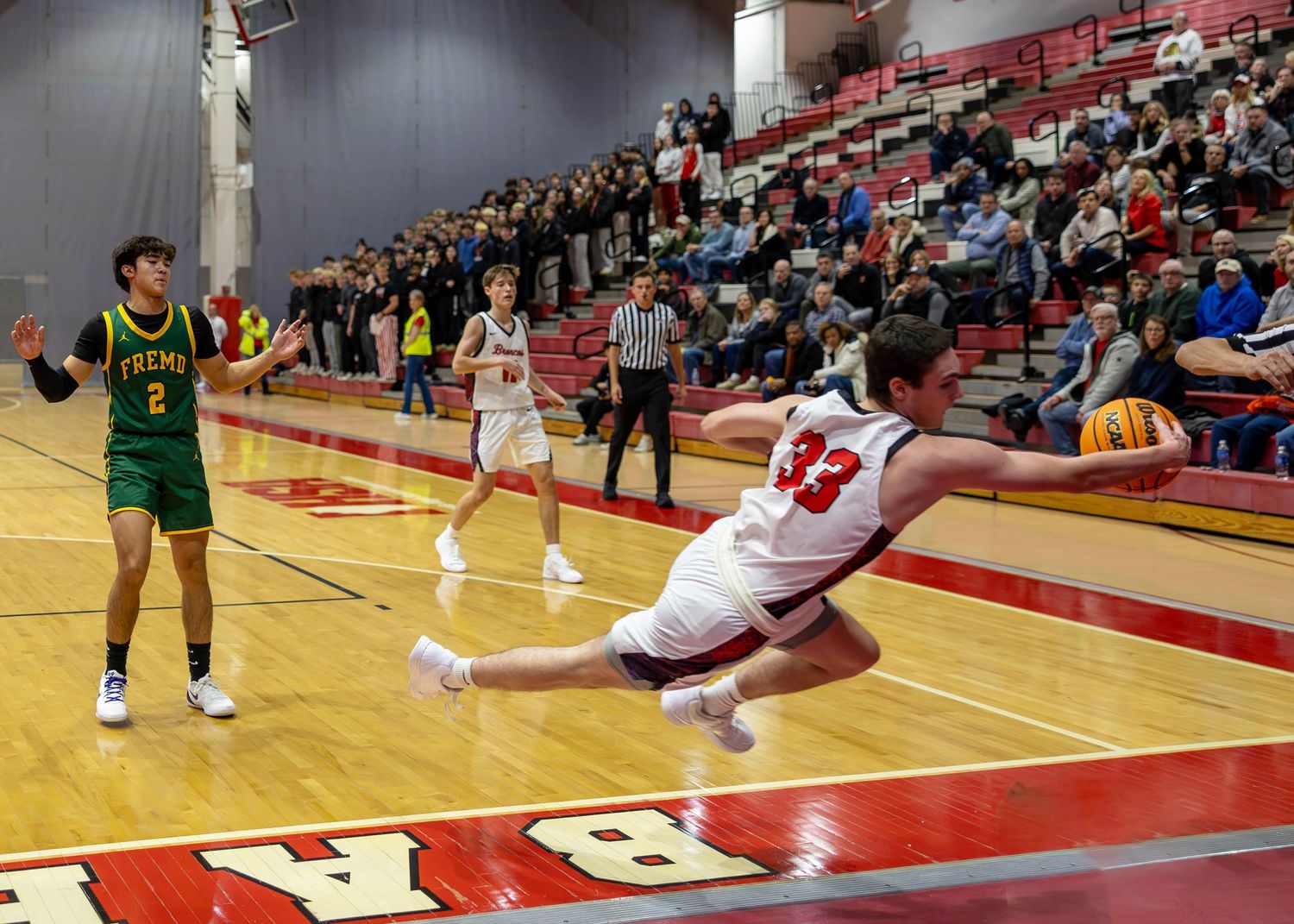 Player #33 dives for the ball during an intense high school basketball game in a packed gymnasium.