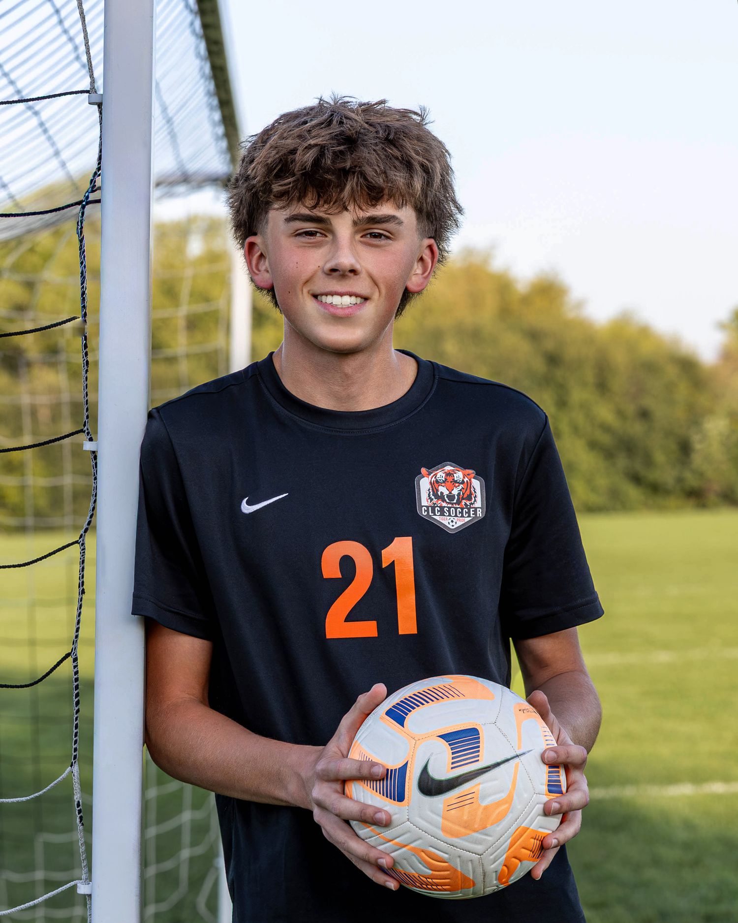 Soccer player in black jersey holds orange ball while posing near goal post on sunny field.