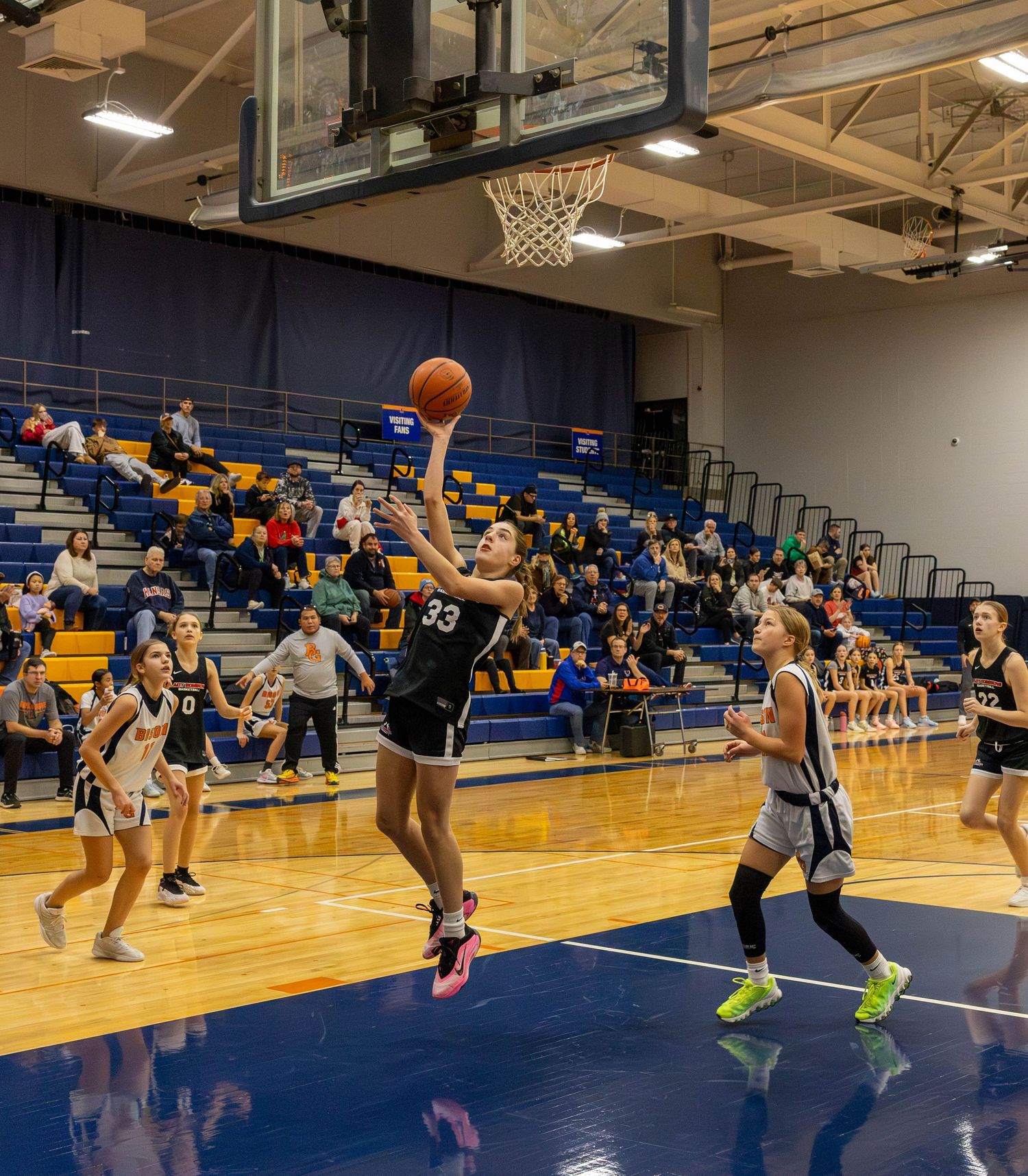 Player #33 in black uniform leaps for a layup during a girls youth basketball game in a packed gymnasium.