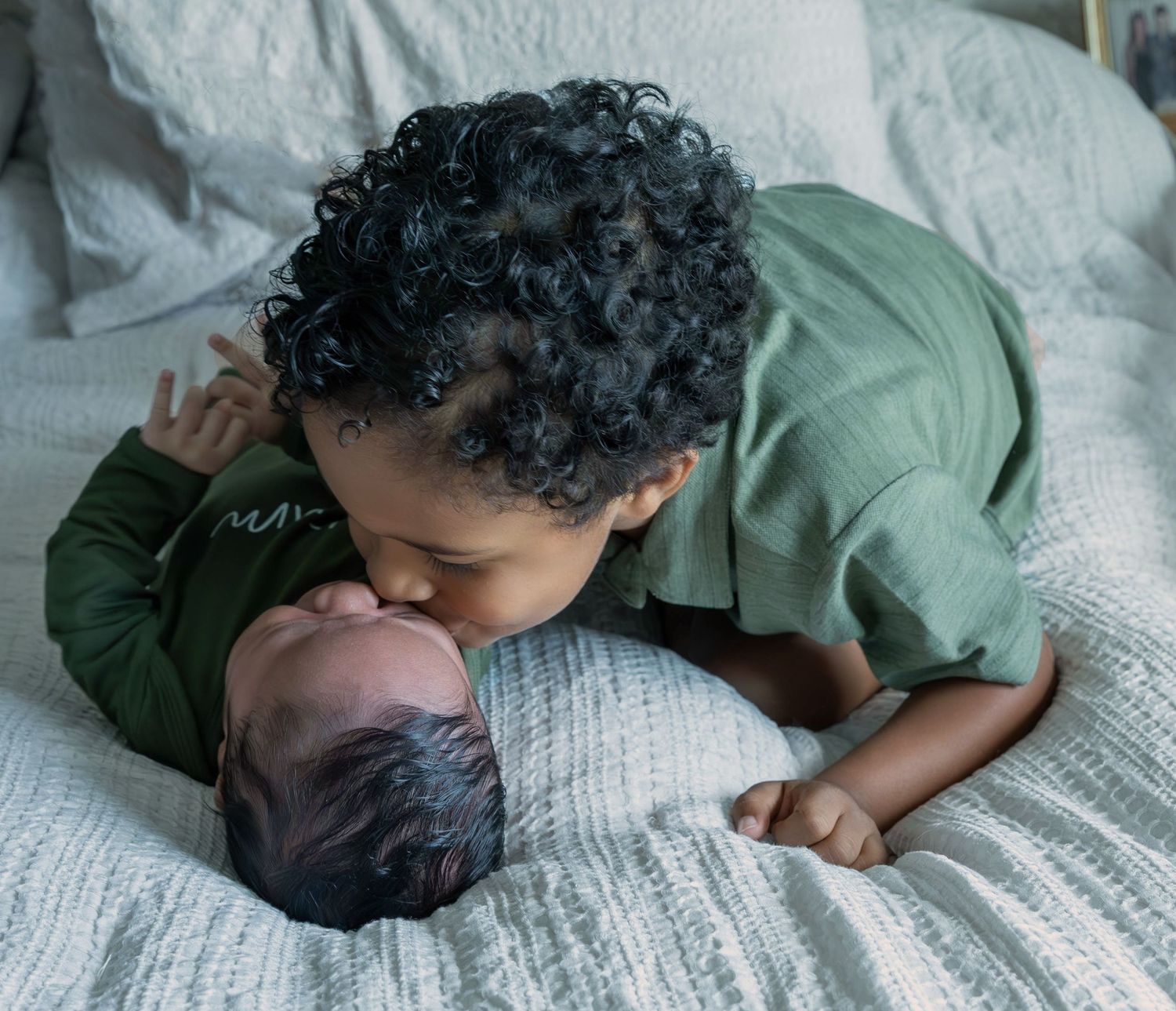 A young toddler with curly hair gently kisses a newborn baby lying on a white bed.