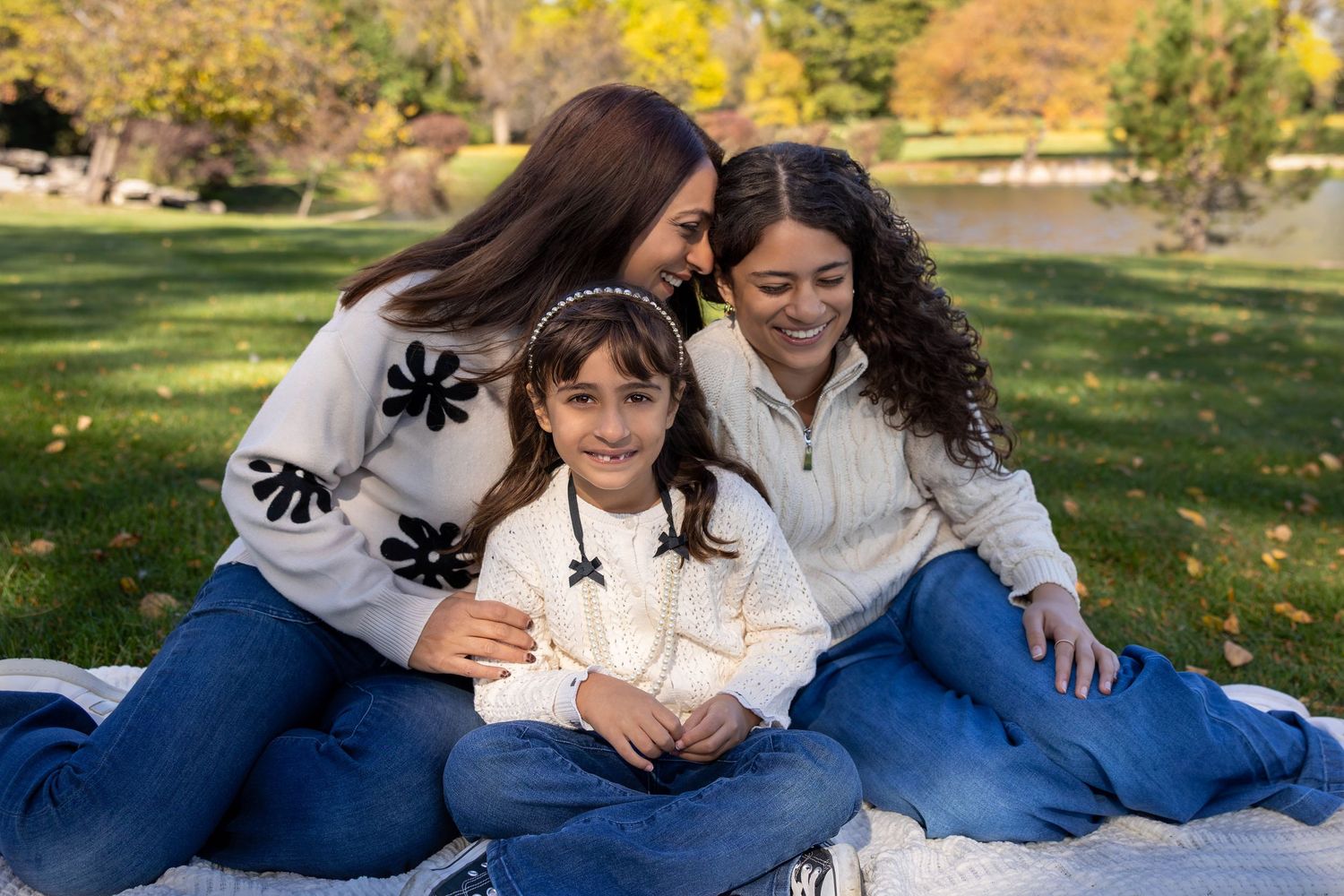 Two women and a young girl smile together on a blanket in a sunny park with autumn foliage.