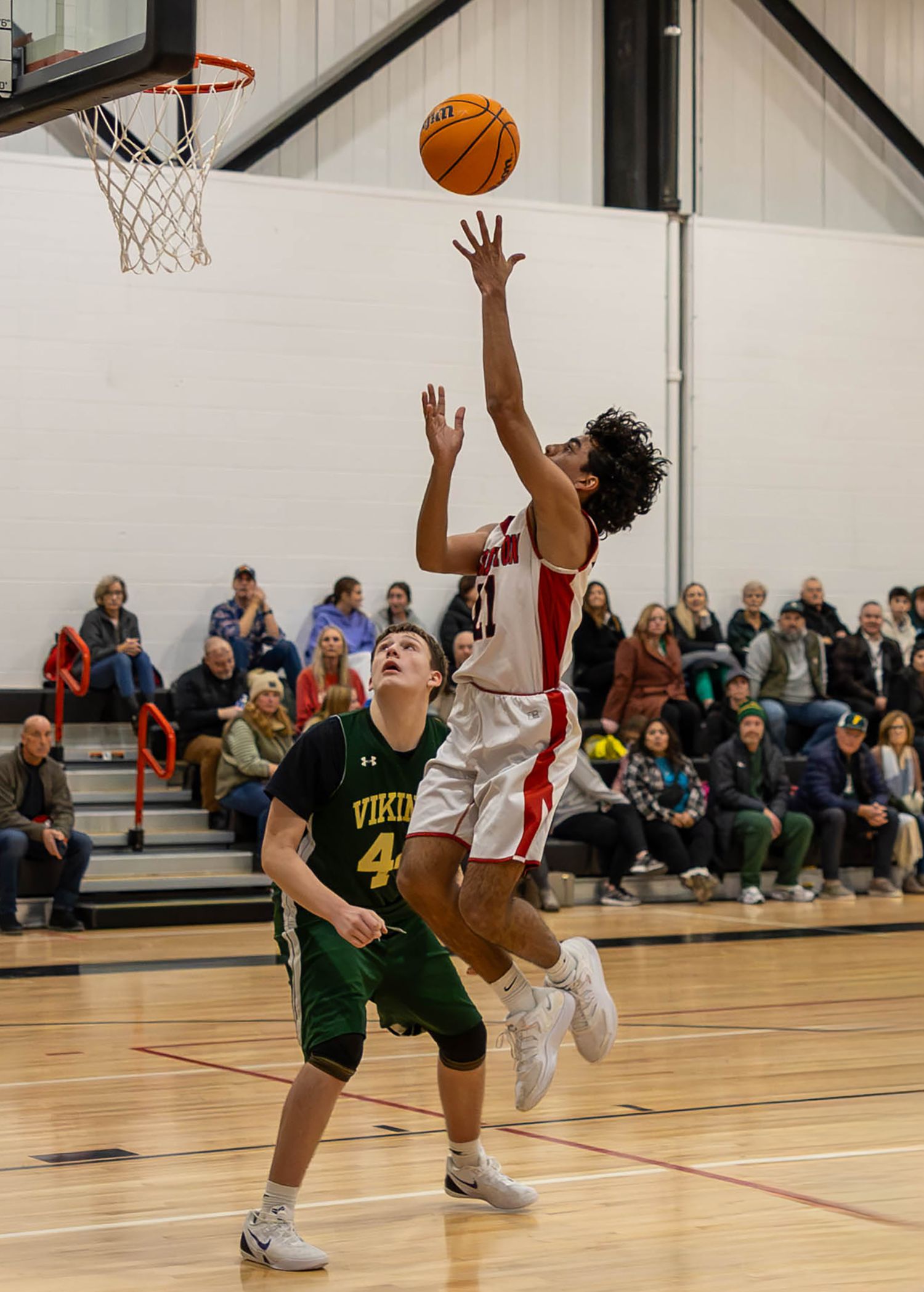 High school basketball player in red uniform drives to the basket past a defender in green Vikings jersey during indoor game.