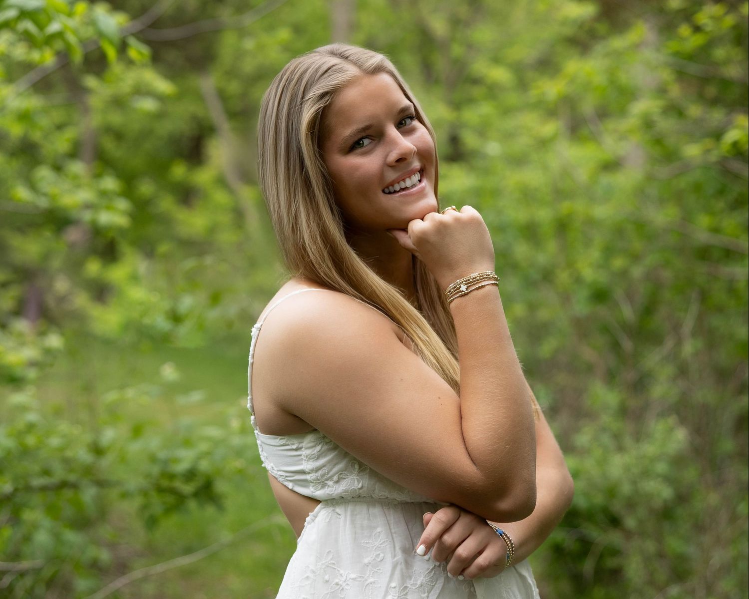 Smiling young woman in white dress posing outdoors with lush green trees in background.