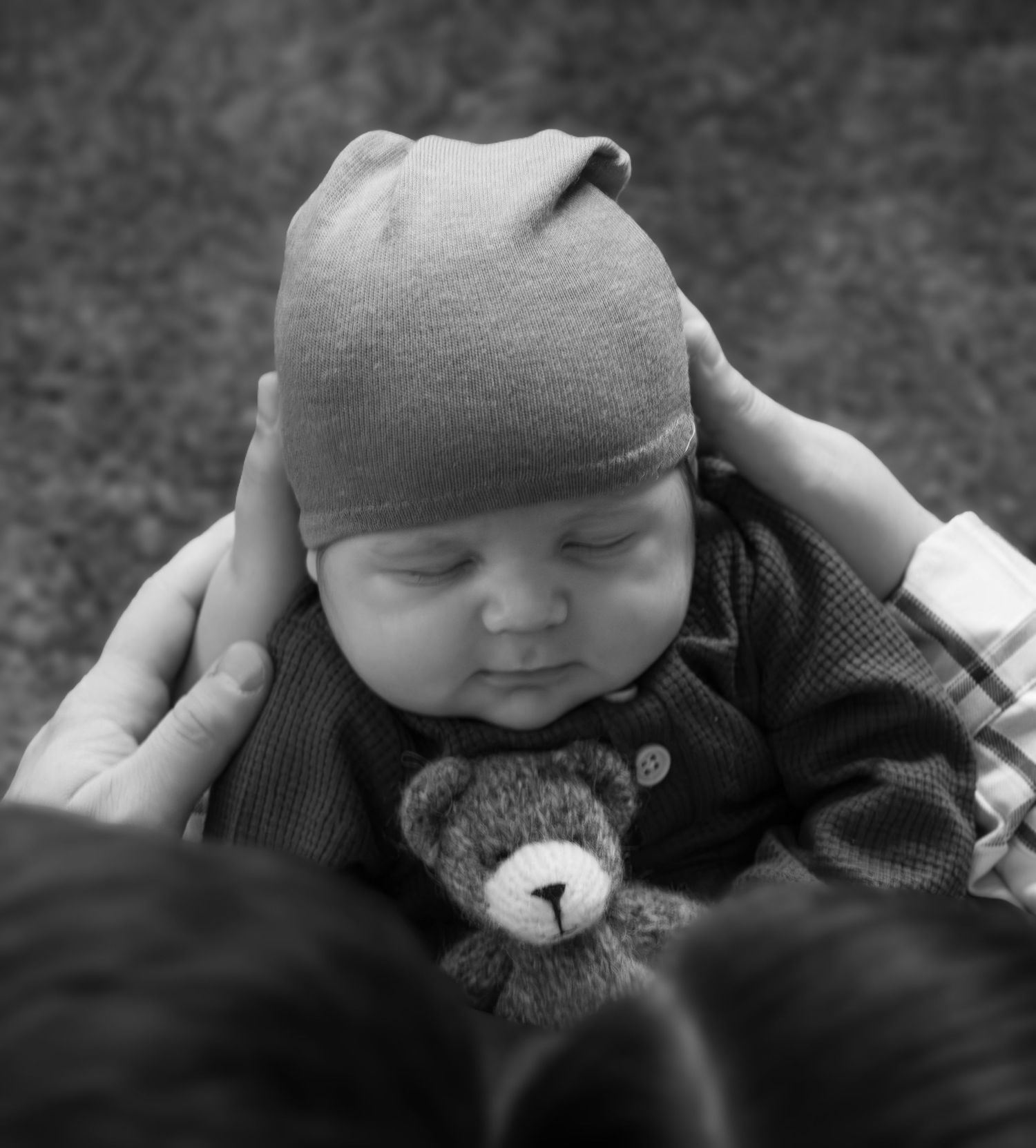 Black and white photo of a sleeping newborn baby wearing a knit hat, cradled in hands, holding a tiny teddy bear.