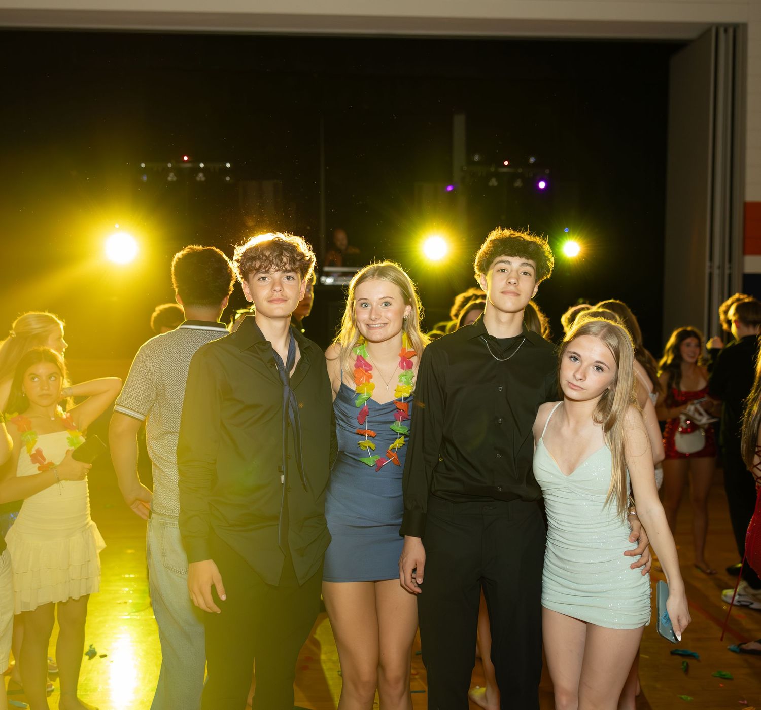 Four teenagers pose together at a school dance, dressed in semi-formal attire under warm yellow stage lighting.