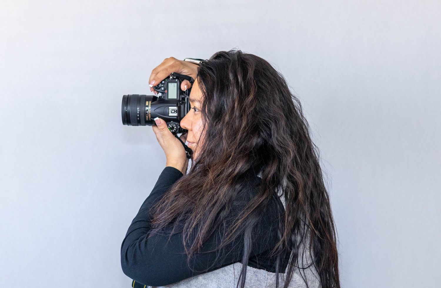 A photographer with long dark hair wearing a black top takes a photo with a professional camera against a gray background.