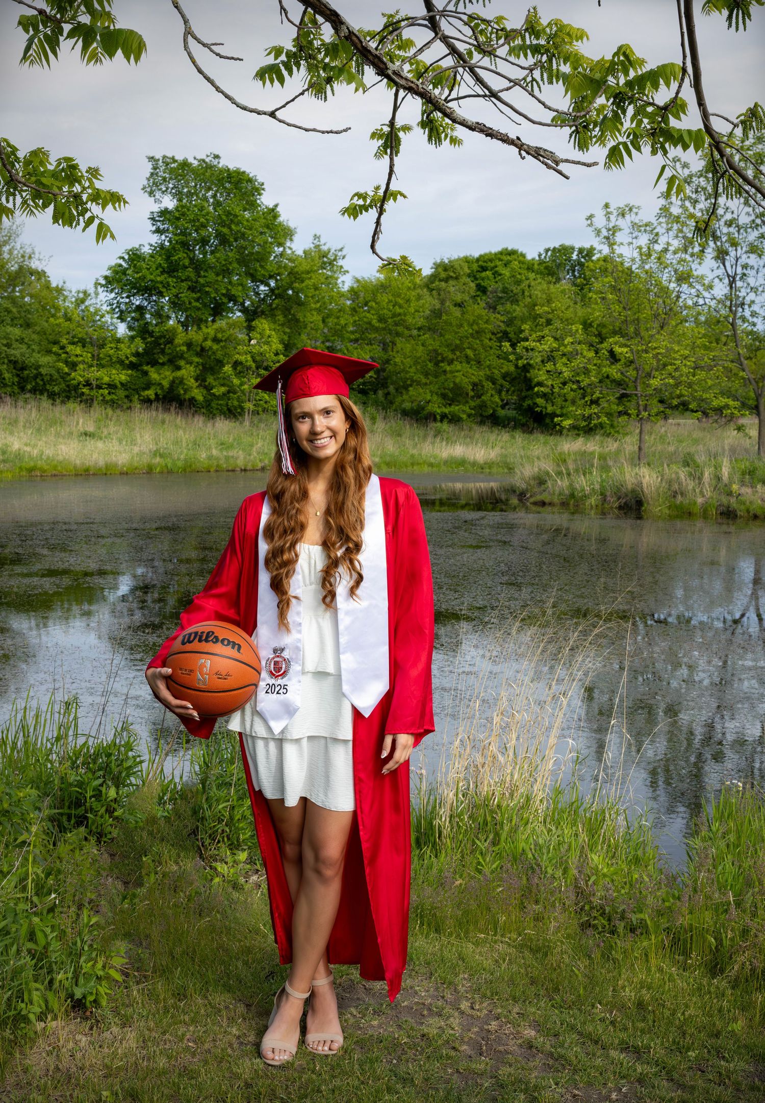 Graduate in red cap and gown holds basketball while standing by a pond in a natural outdoor setting.