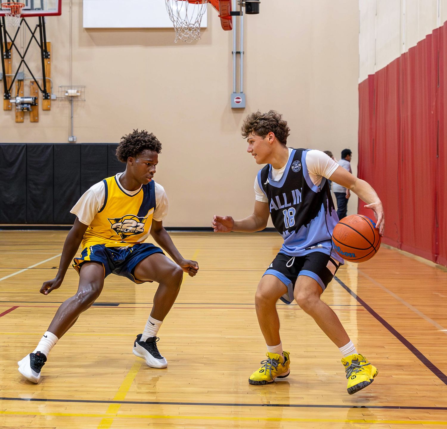 Two teen basketball players compete in an indoor gym, one dribbling in a blue jersey while the other defends in yellow.