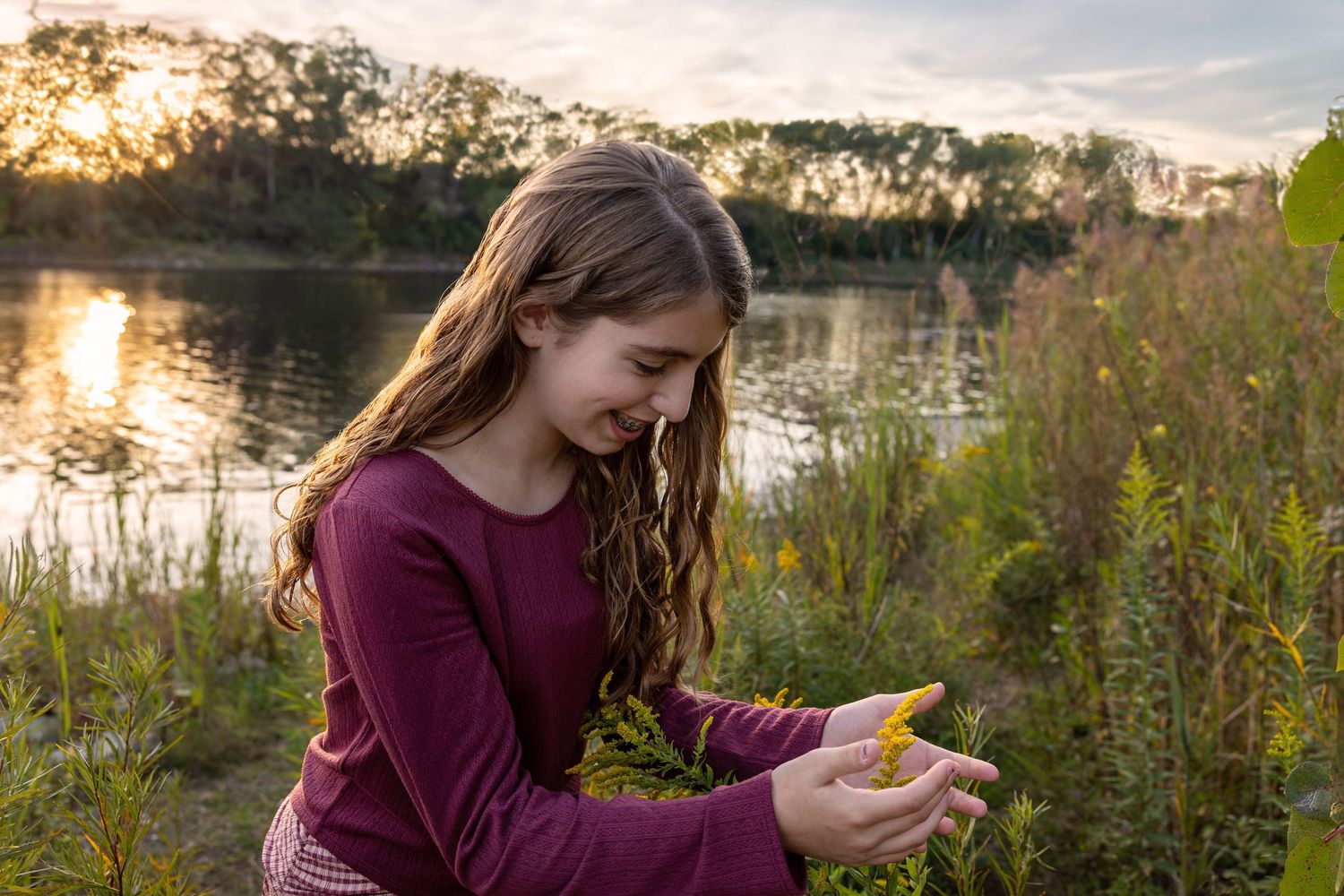 A person in a burgundy sweater examines a small object by a lake during golden hour sunset.
