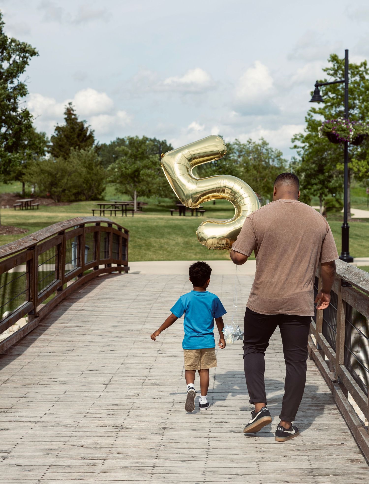 Father and young son walking on a bridge holding a large gold number 5 balloon in a park.