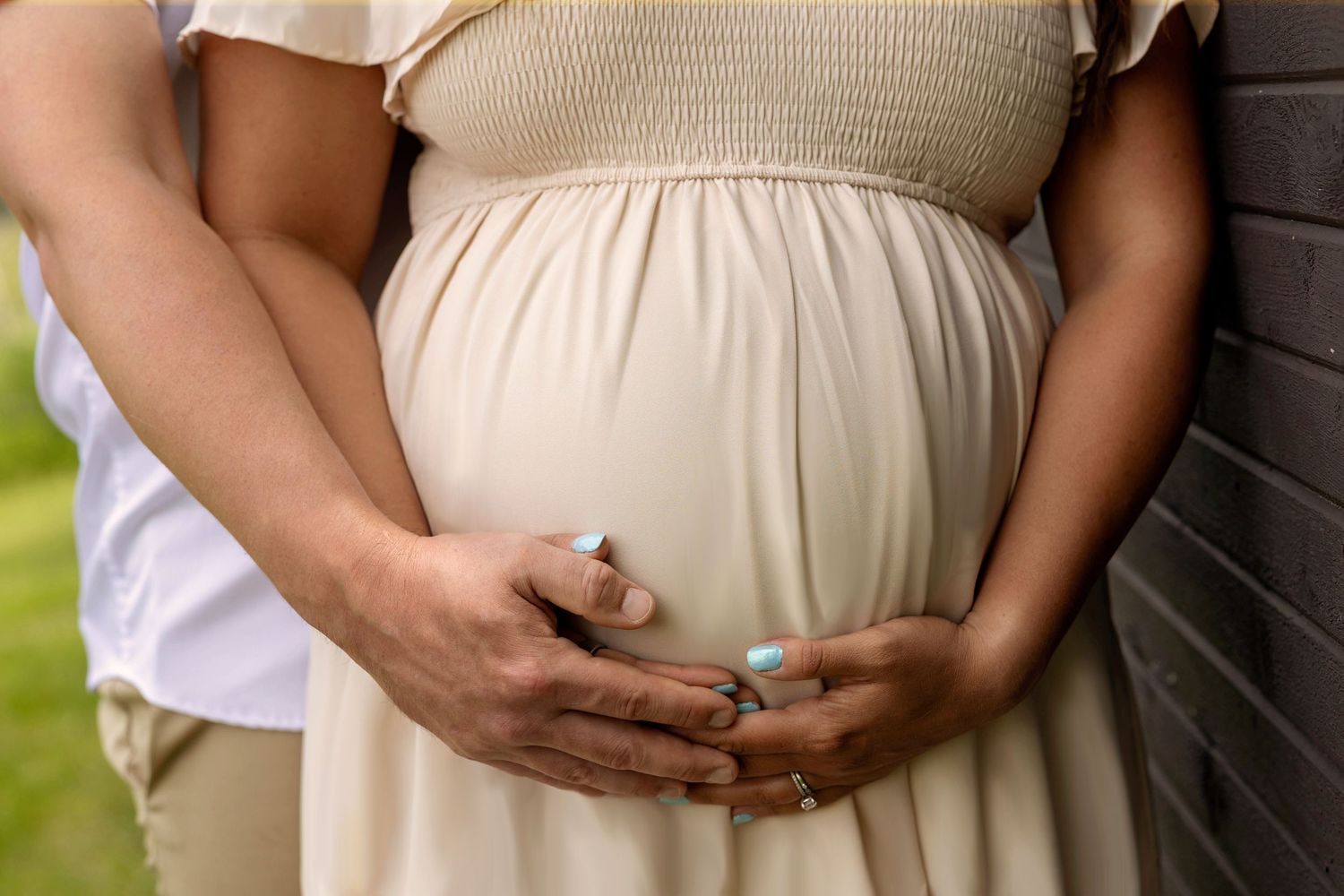 Pregnant woman in beige dress cradling baby bump with partner's hands, maternity photography outdoors.