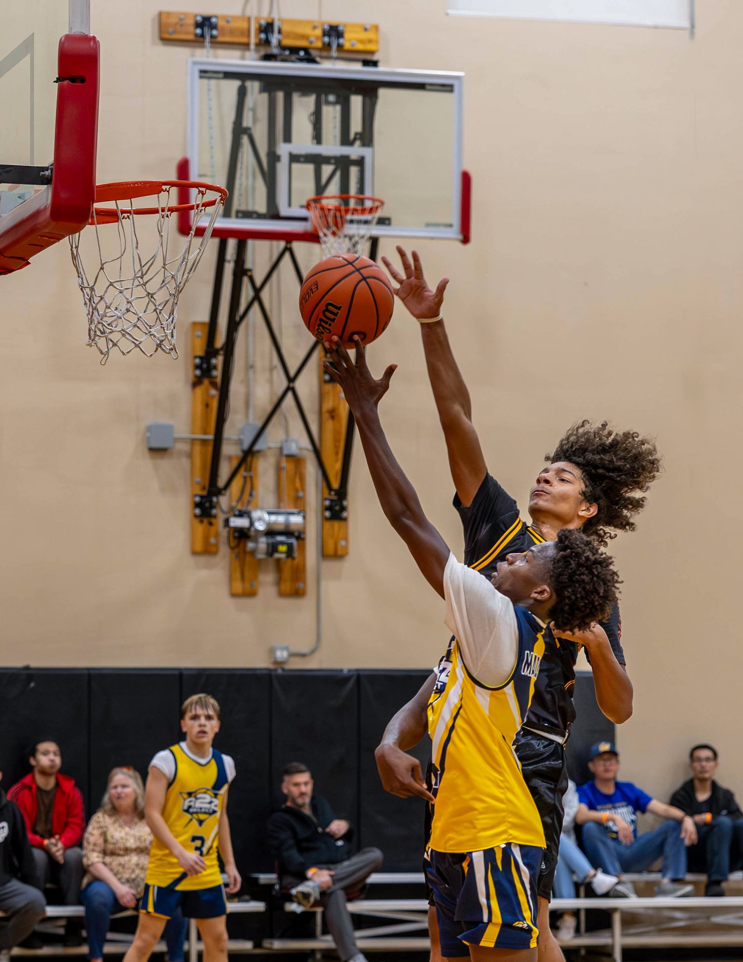 Two basketball players battle for a rebound near the hoop during an indoor youth basketball game.