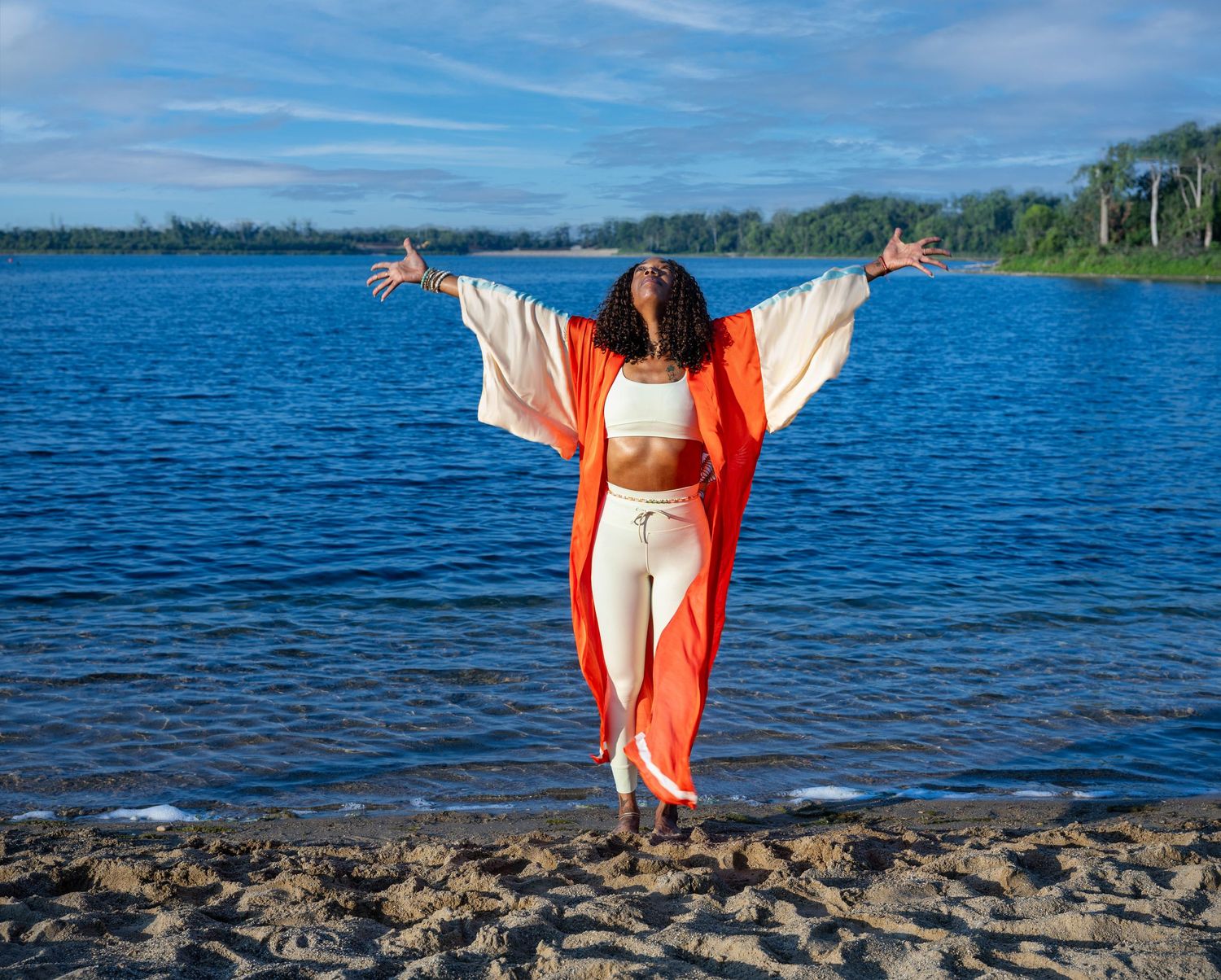Woman in orange kimono and cream outfit stands arms outstretched by a blue lake on a sunny day.