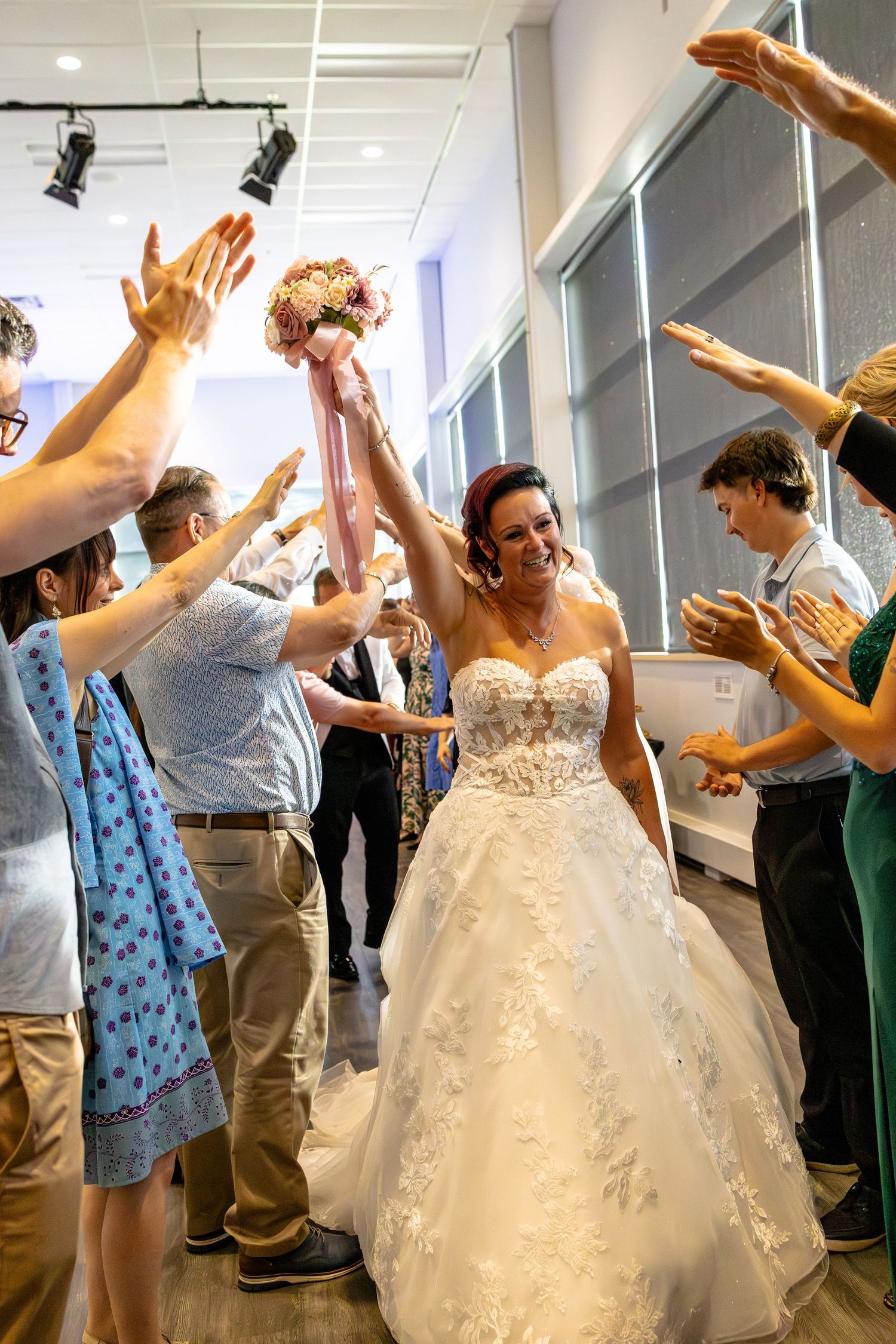 Bride in white lace ball gown walks through cheering guests raising hands, holding bouquet aloft at wedding reception.