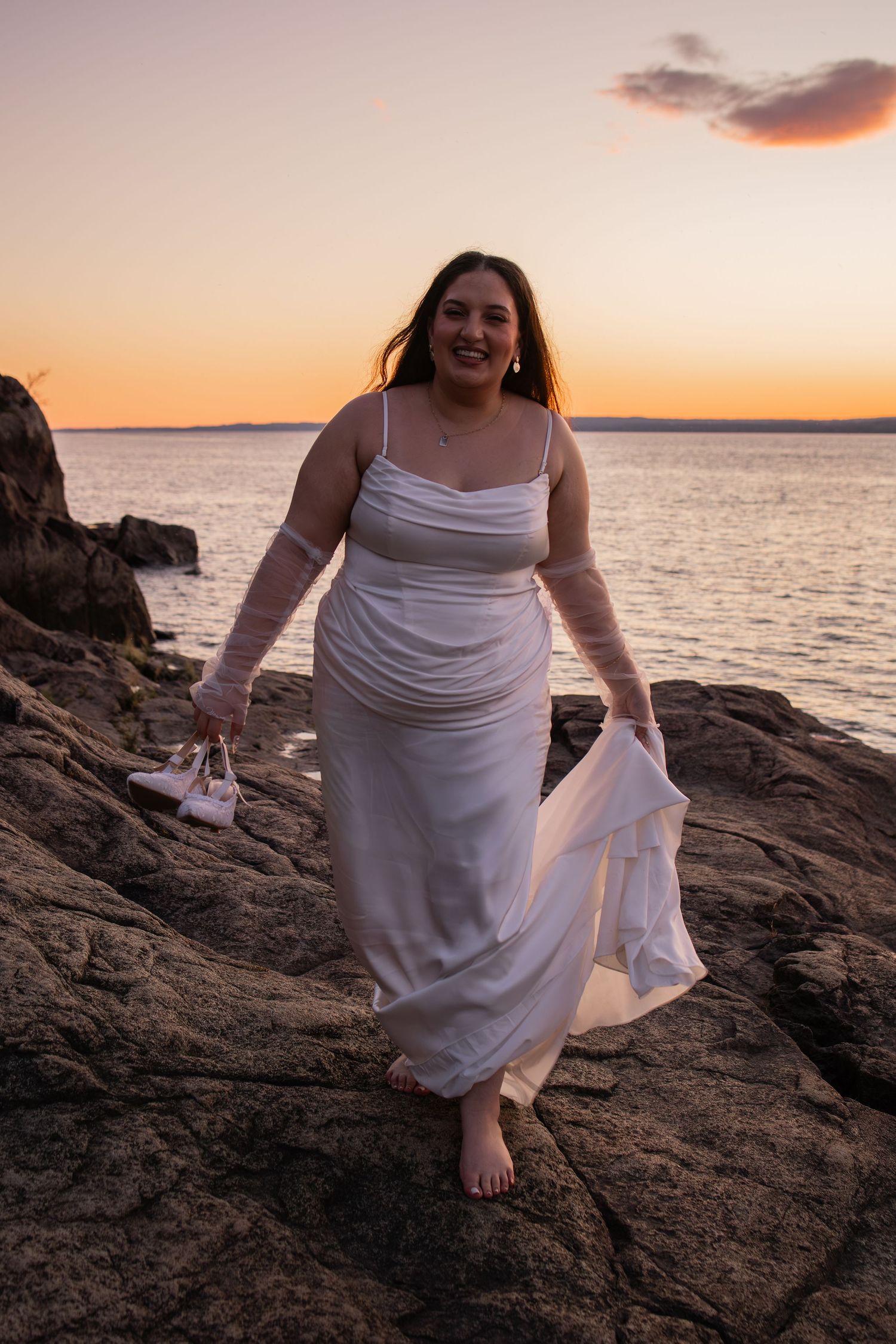 Woman in white dress walking barefoot on rocky shore at sunset, holding shoes and flowing fabric.