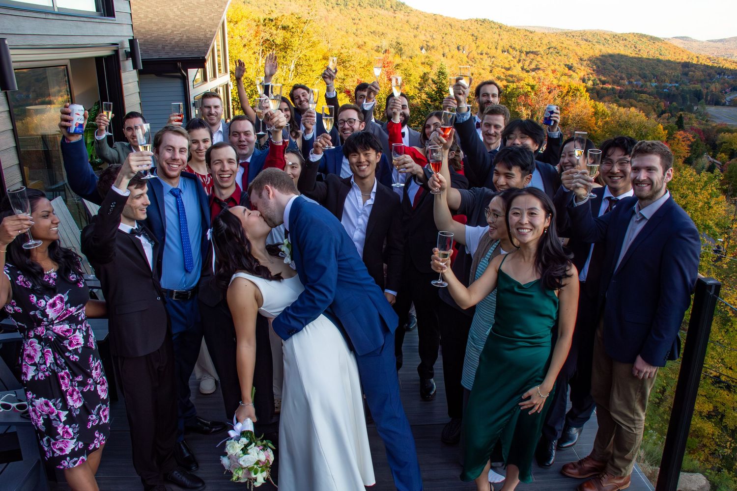 Bride and groom kiss surrounded by cheering wedding guests on outdoor terrace with fall foliage.