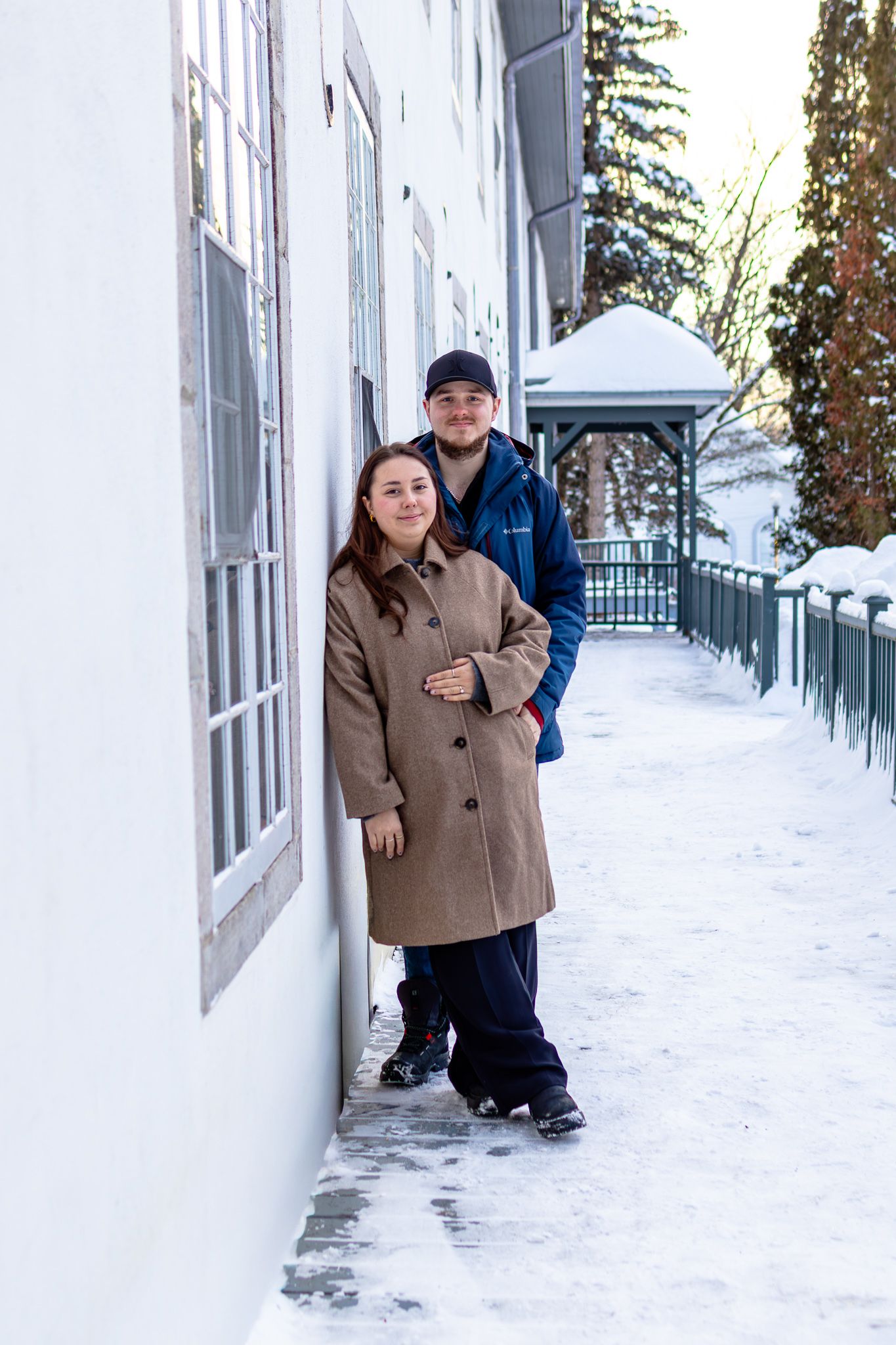 Couple in winter coats leaning against white building wall on snowy day outdoors.