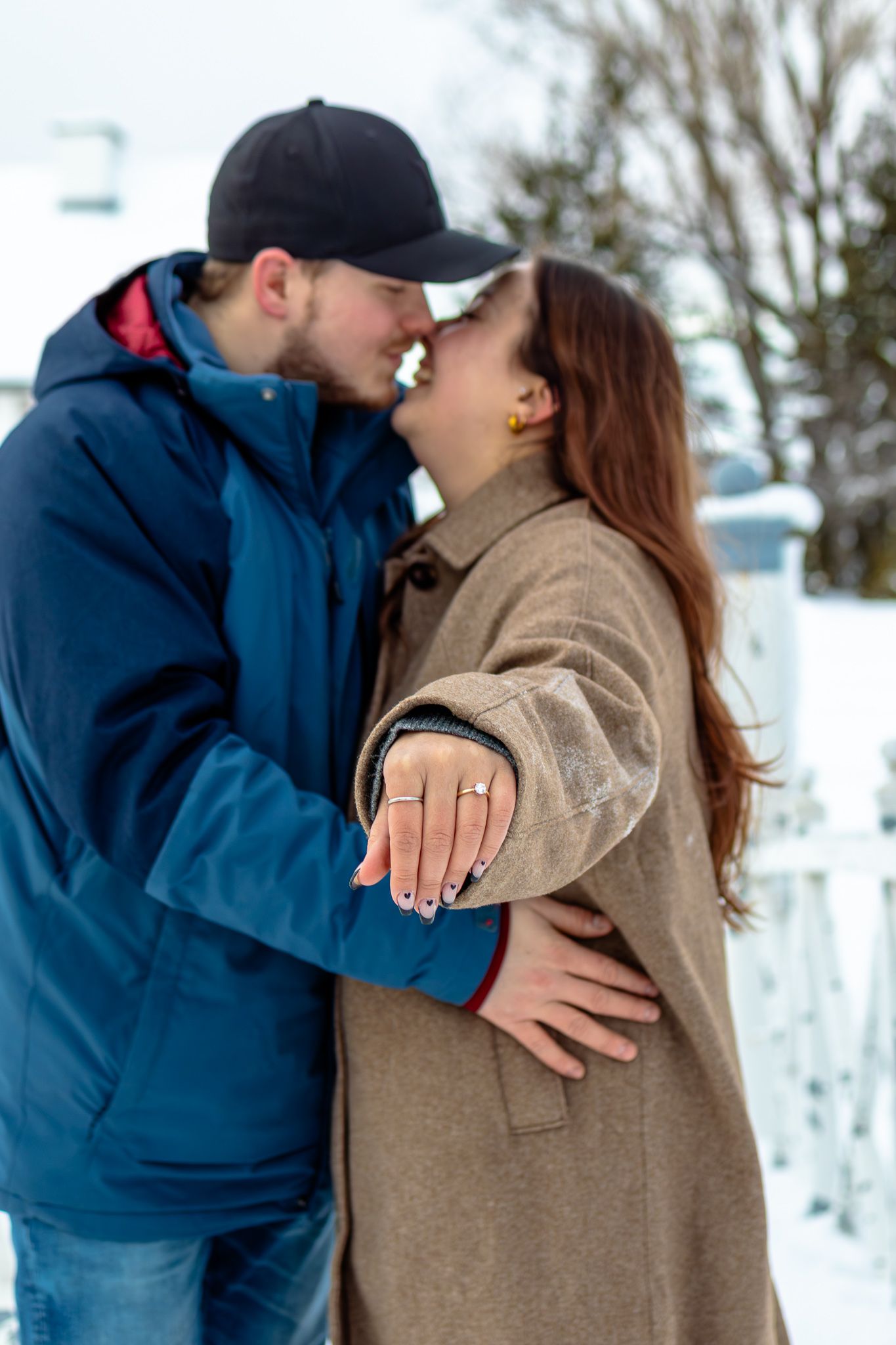 Engaged couple sharing a kiss outdoors in winter, woman showing off diamond engagement ring on her outstretched hand.