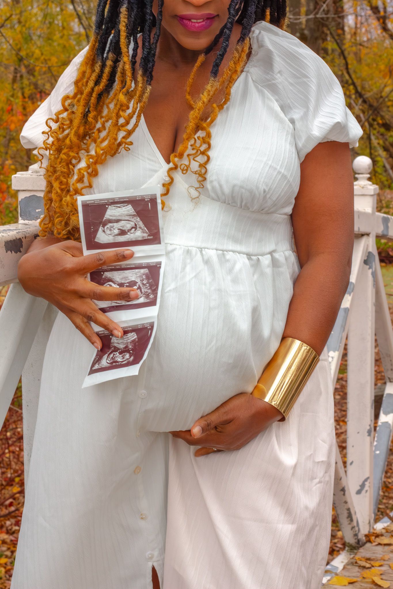 Pregnant woman in white dress holding ultrasound photos against her baby bump in an autumn park.