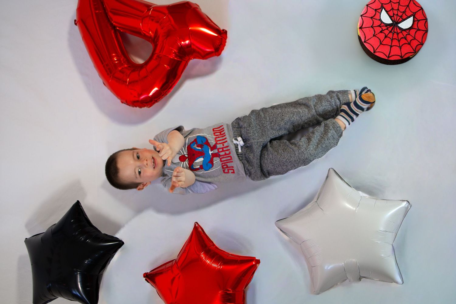 Baby boy in Spiderman outfit lying among star and number balloons with a Spiderman cake nearby.