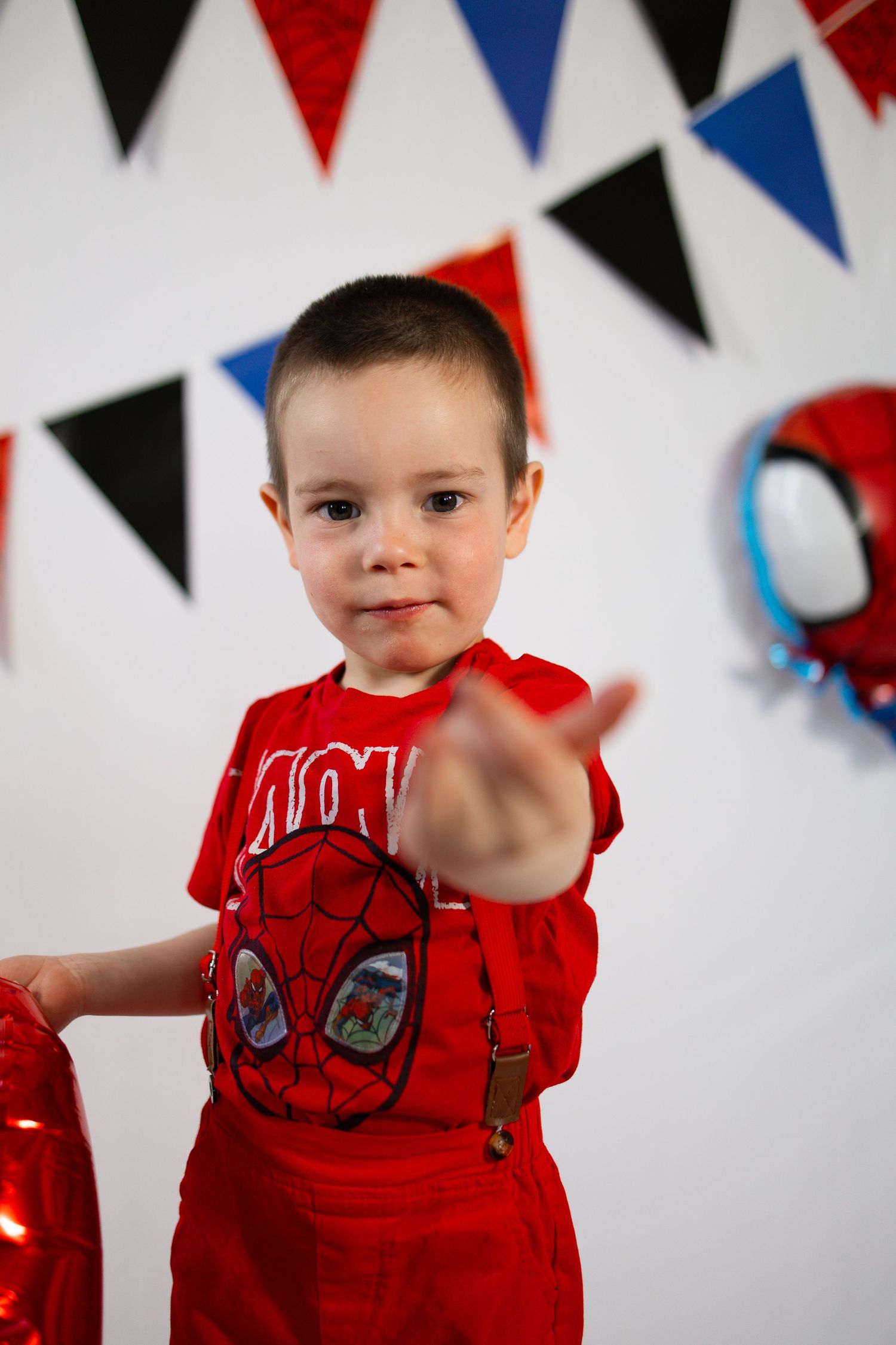 A young boy in a red Spiderman shirt and suspenders points at the camera at a Spiderman-themed birthday party.