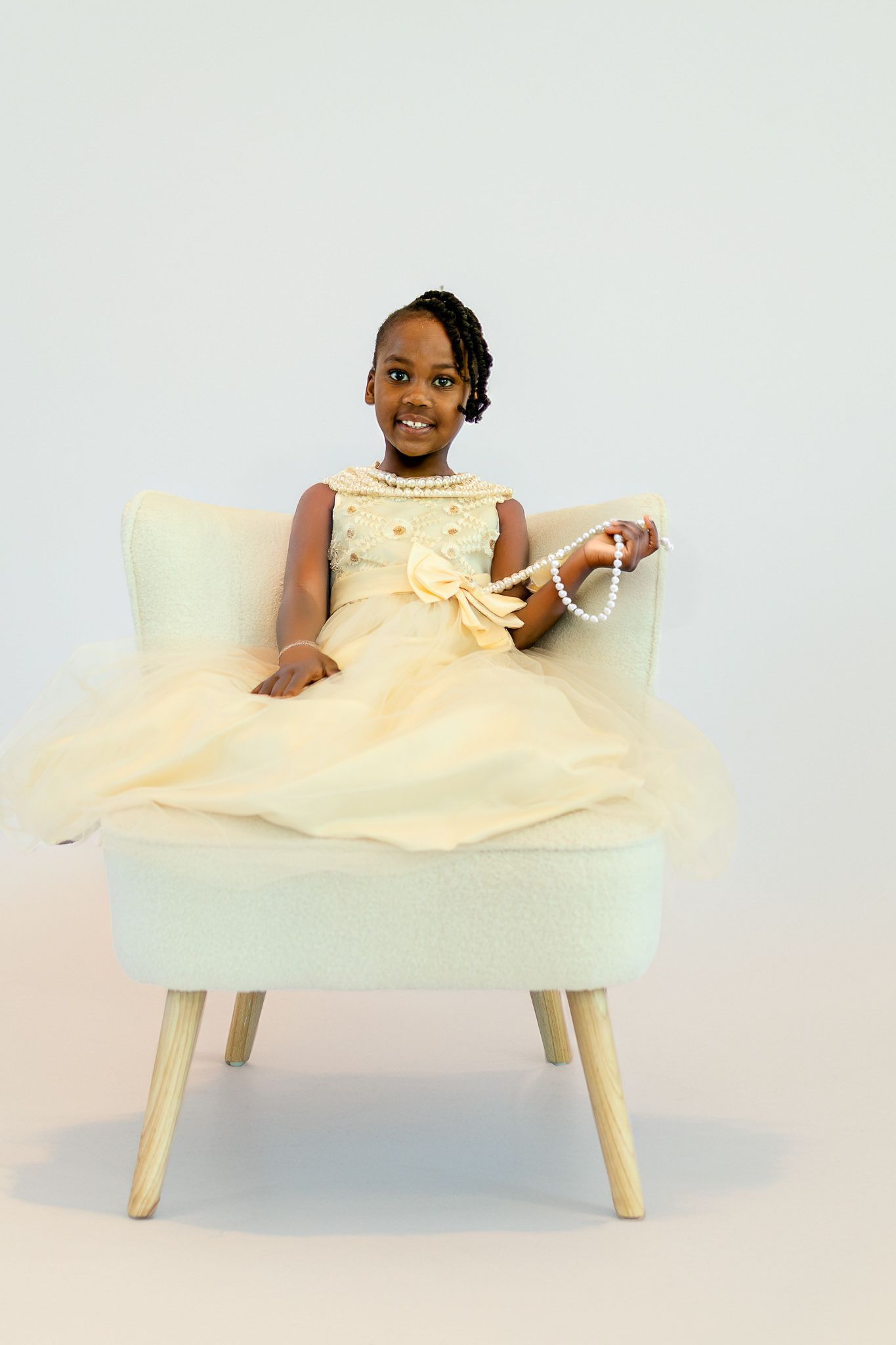 Young girl in yellow dress sitting in white chair holding pearl necklace, smiling at camera.