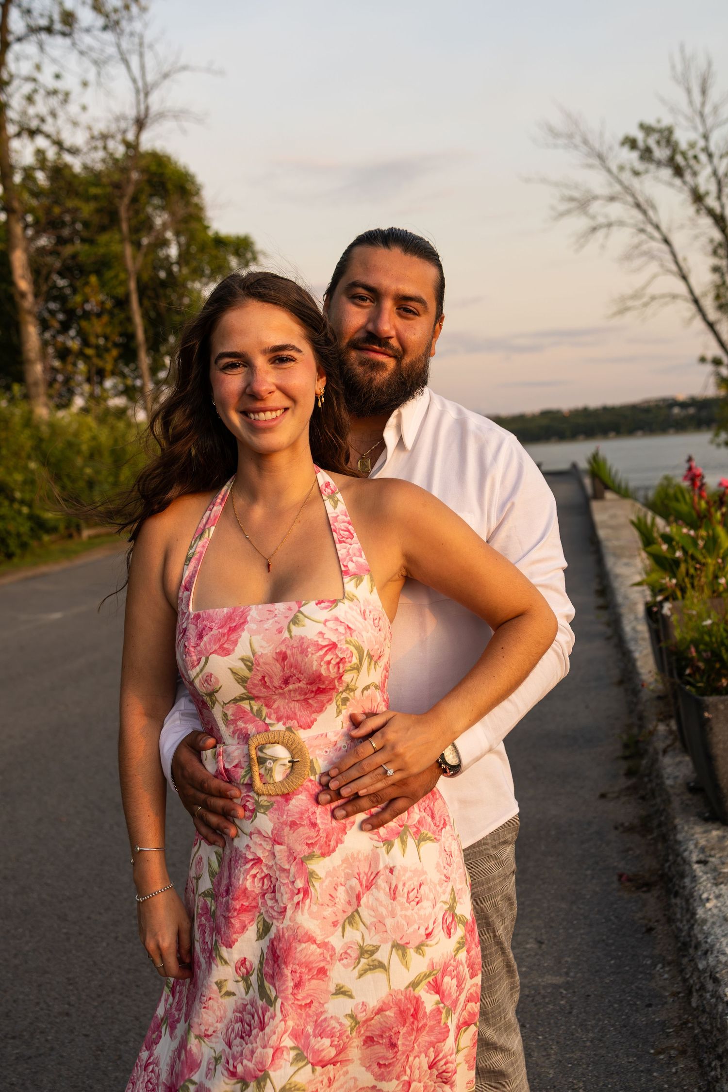 Couple posing outdoors at sunset, woman in floral dress, man in white shirt, waterfront road background.
