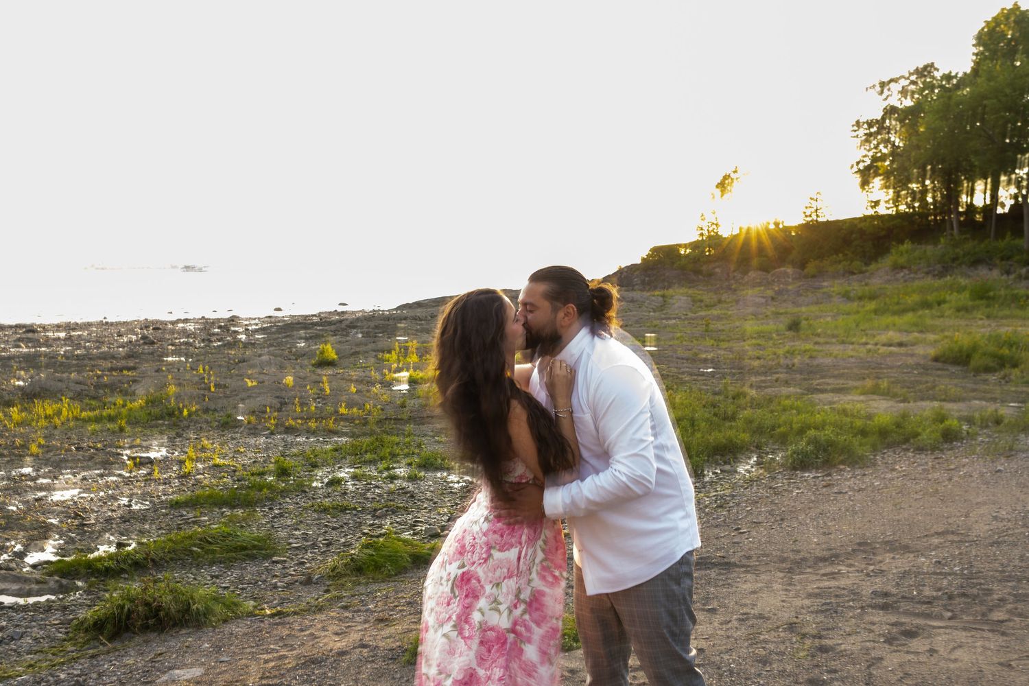 Couple sharing a romantic kiss at sunset on a rocky shoreline, woman in floral dress, man in white shirt.