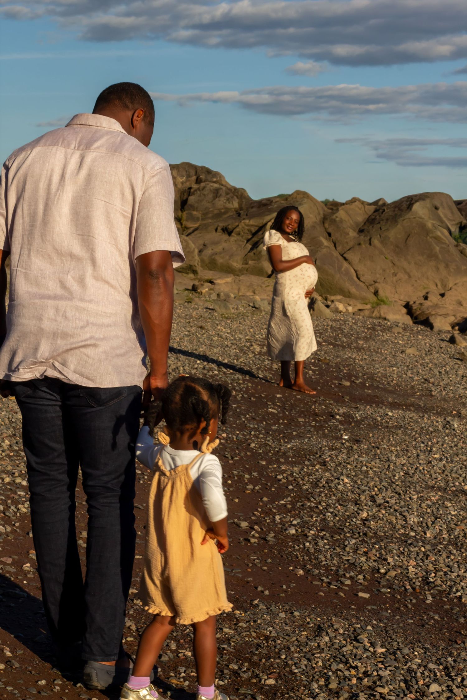 Black father and toddler daughter walking toward pregnant mother on pebble beach at sunset.