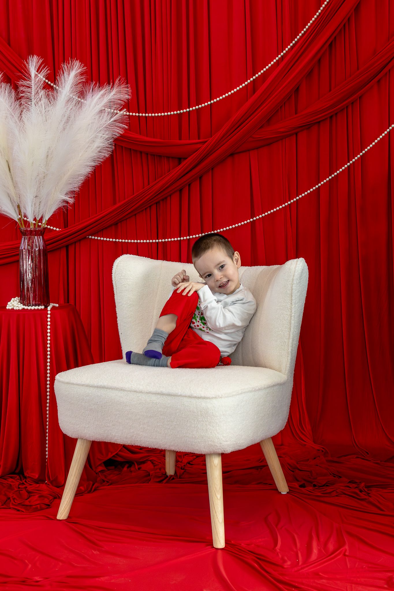 Young boy sitting in white armchair holding red item against decorative red curtain backdrop.
