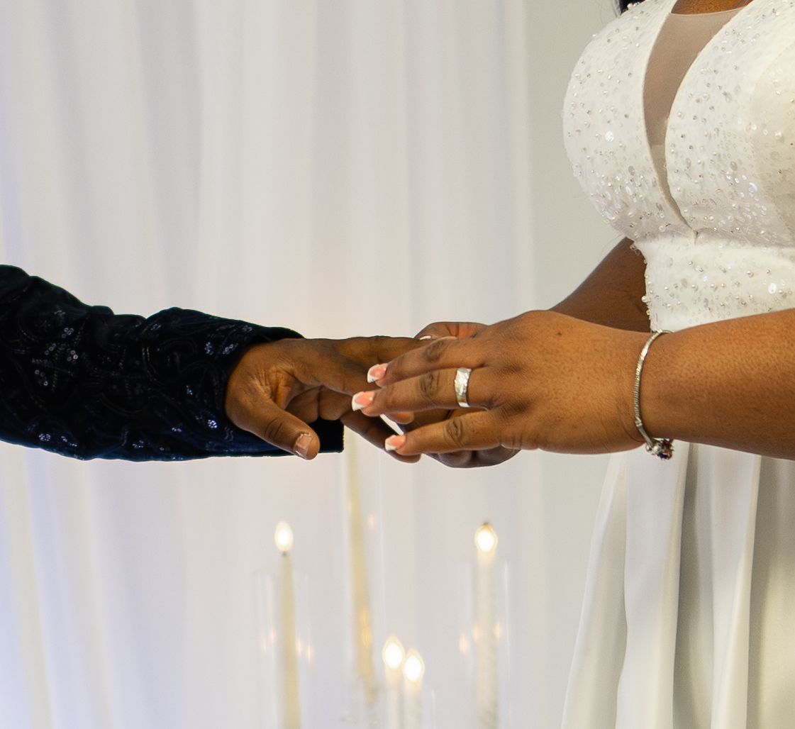 A bride in white lace dress places a gold wedding ring on groom's finger during wedding ceremony with candles.