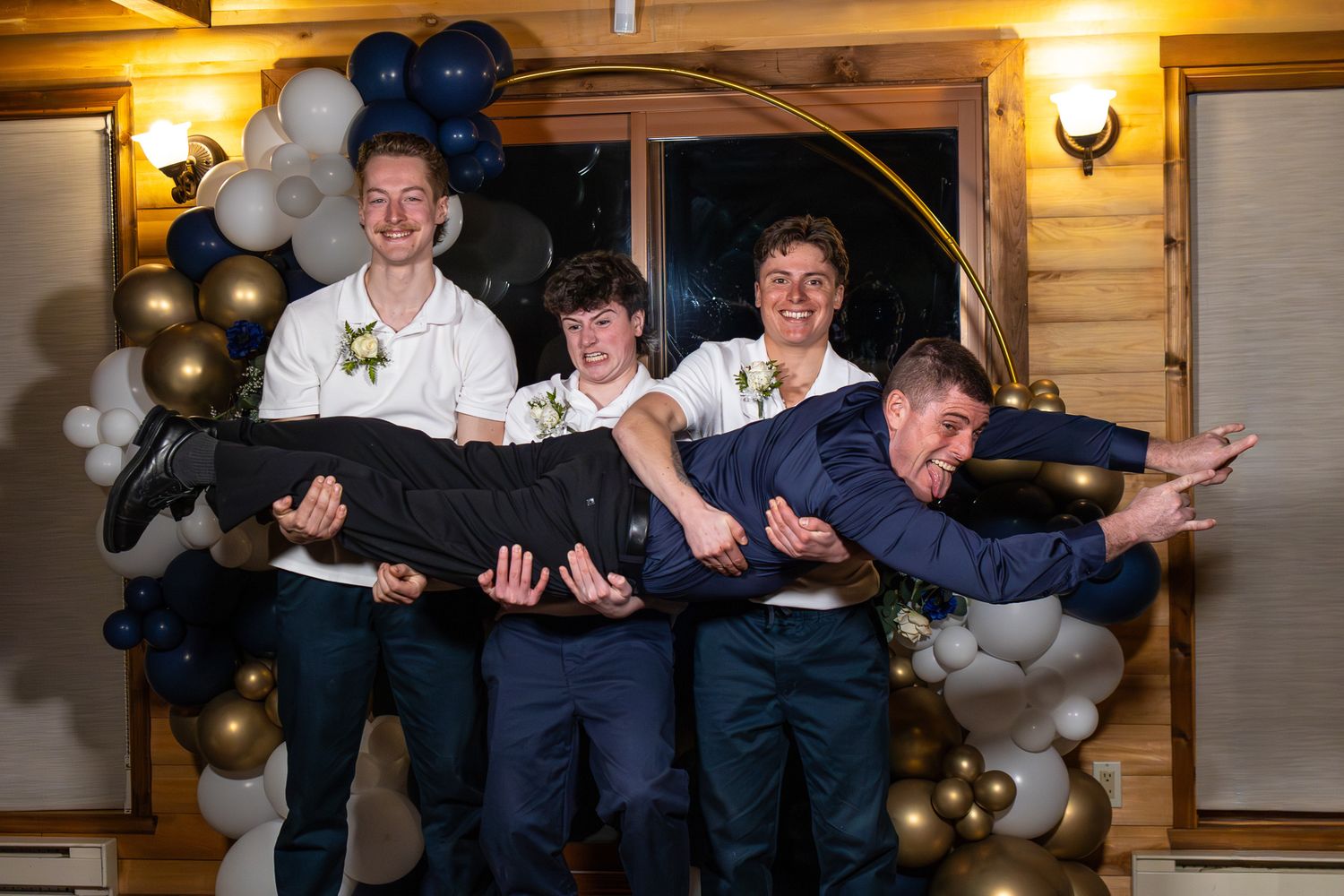 Wedding party playfully lifting groom horizontally in front of navy blue and gold balloon decorations.