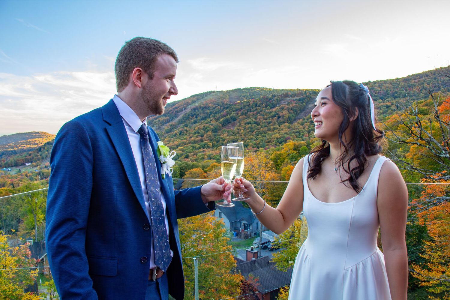 Bride and groom toast with champagne glasses overlooking colorful autumn mountain scenery.